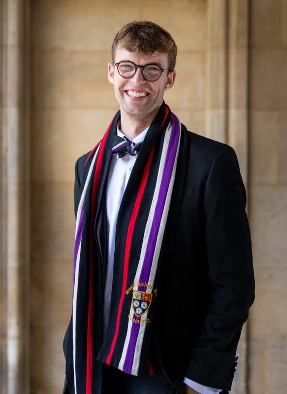 Young man smiling, wearing glasses, a tuxedo, and a graduation scarf with the university emblem, standing outside with a stone building in the background.