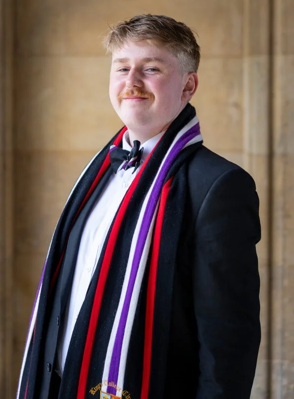 A young man dressed in formal attire, including a tuxedo and bow tie, wearing multiple colorful scarves, standing in front of a stone wall, smiling at the camera.