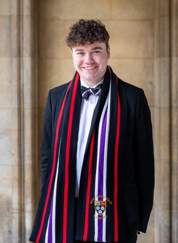 A young man smiling and wearing a formal suit with a striped scarf and a bow tie, standing in front of a stone wall.