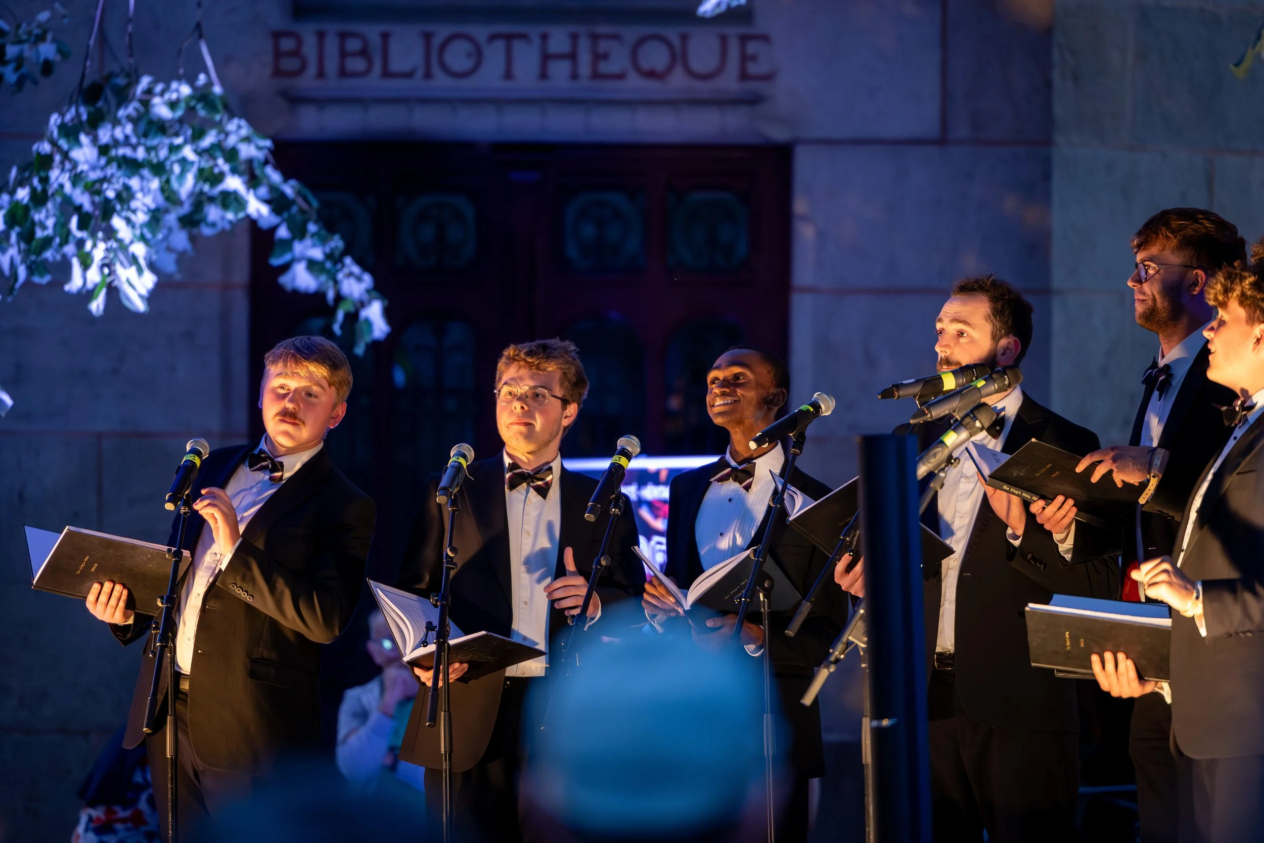 Group of six men in tuxedos singing with sheet music and microphones in an outdoor performance at night in front of a stone building with the sign 'BIBLIOTHEQUE'.