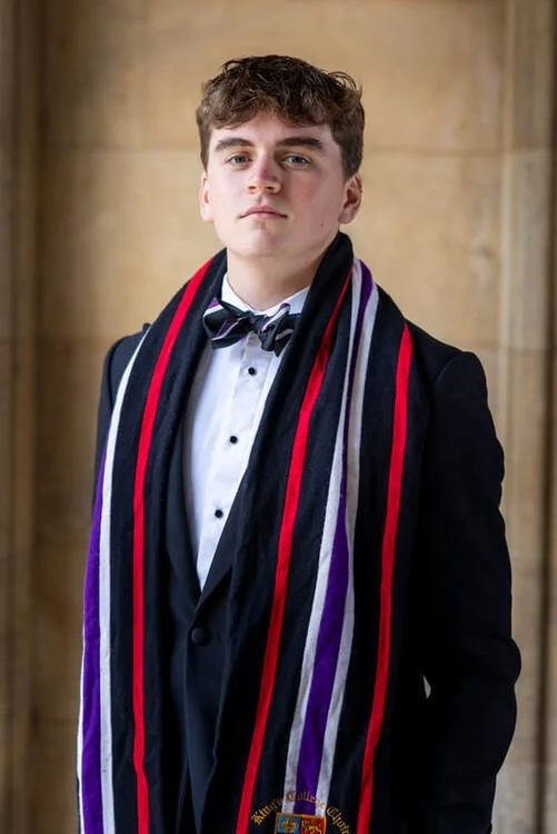 Young man in a black tuxedo with a bow tie, wearing a colorful sash, standing indoors with a stone wall background.