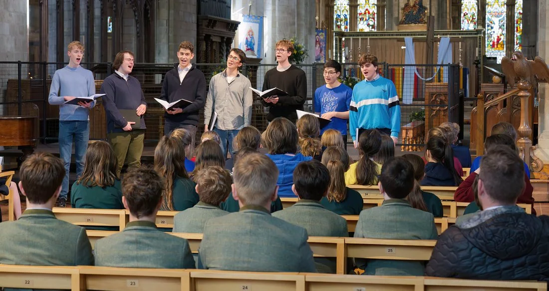 Young boys singing in a choir in front of seated children inside a church with stained glass windows.