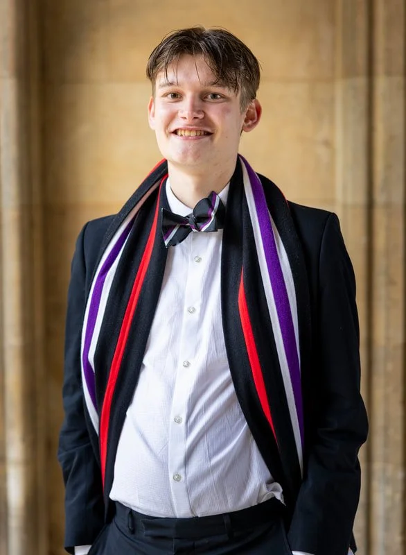 Young man in a tuxedo with a rainbow-colored scarf and bow tie, smiling, standing indoors with wooden background.