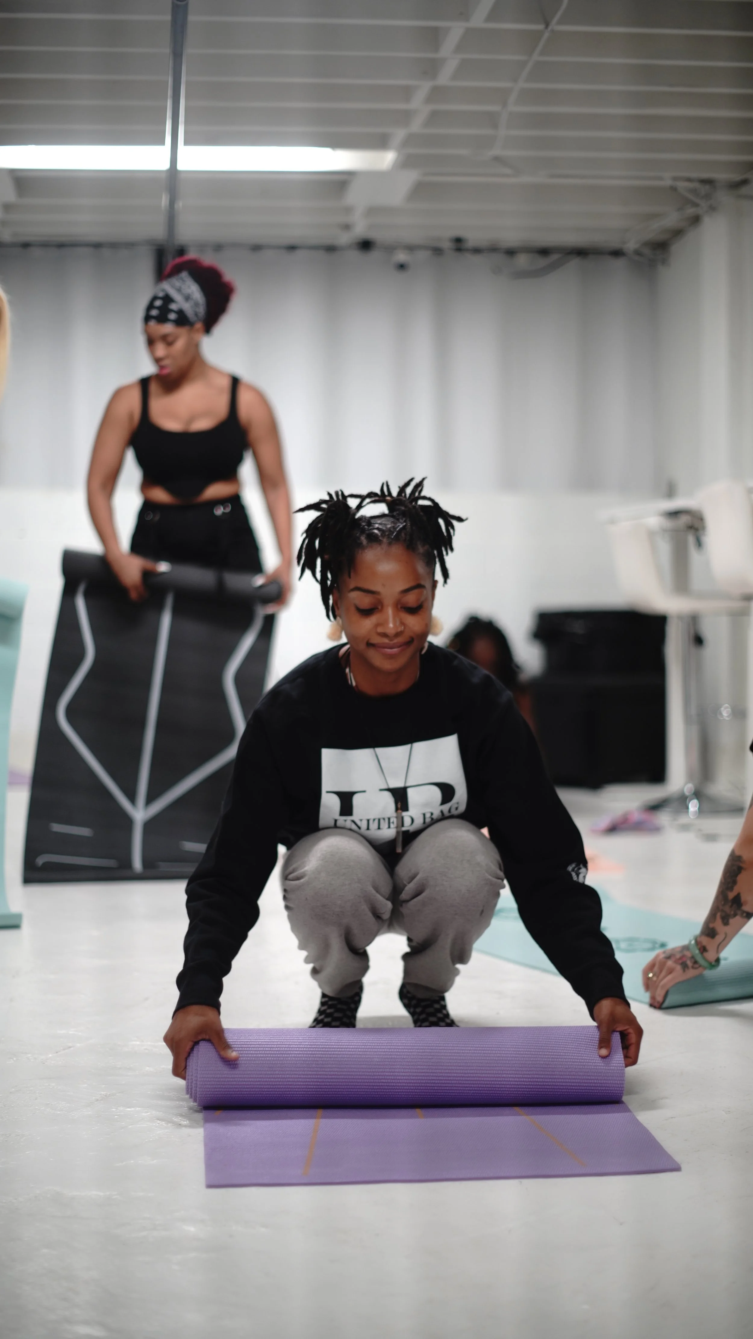 Women practicing yoga in a studio, one is kneeling and rolling out a purple yoga mat, others are in the background preparing for practice.