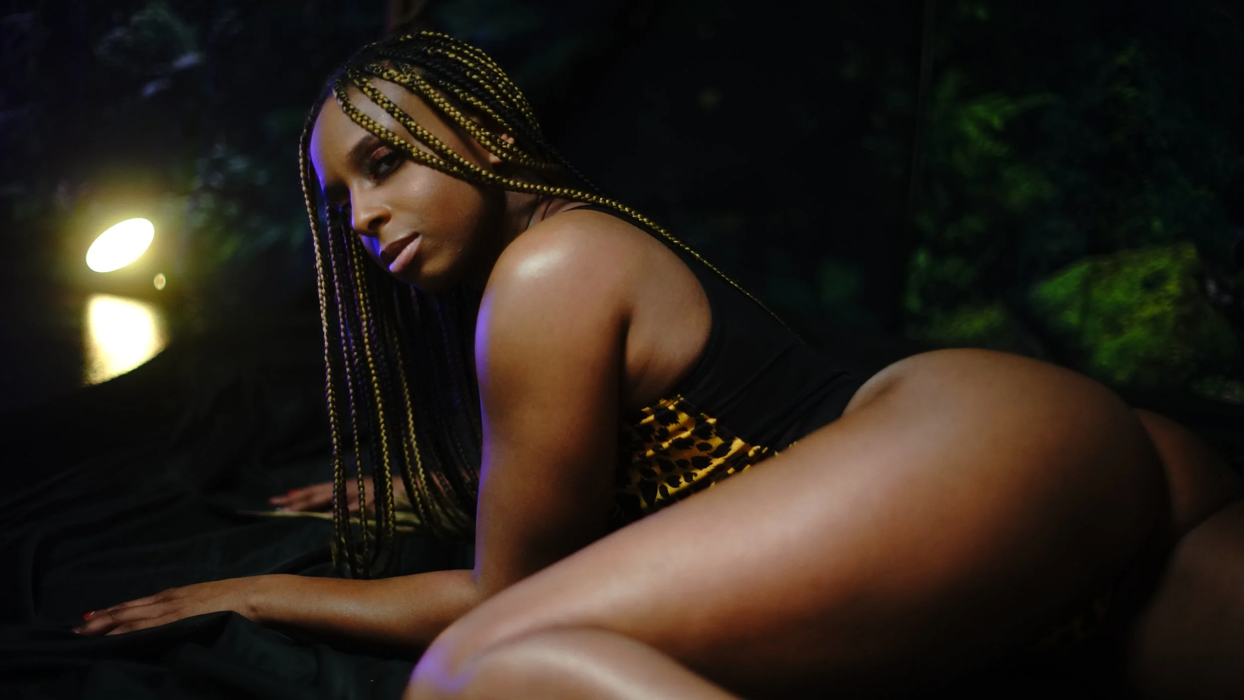 A woman with braided hair and leopard print clothing poses on her hands and knees in a dark, outdoor setting illuminated by a bright light in the background.