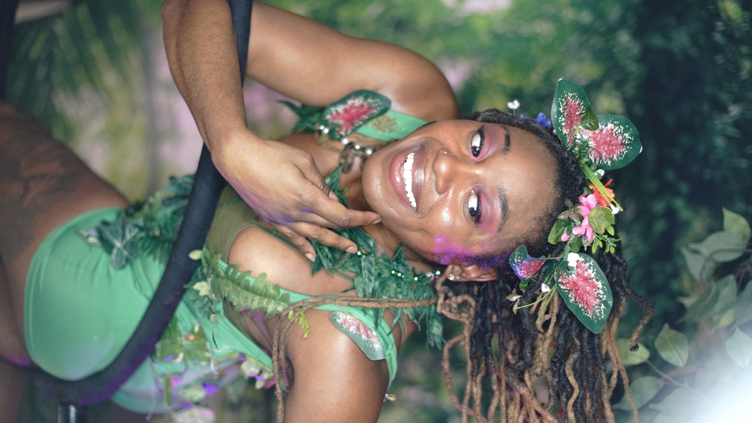 A woman dressed in green with leaf and flower accessories, smiling and posing outdoors in a lush, green setting.
