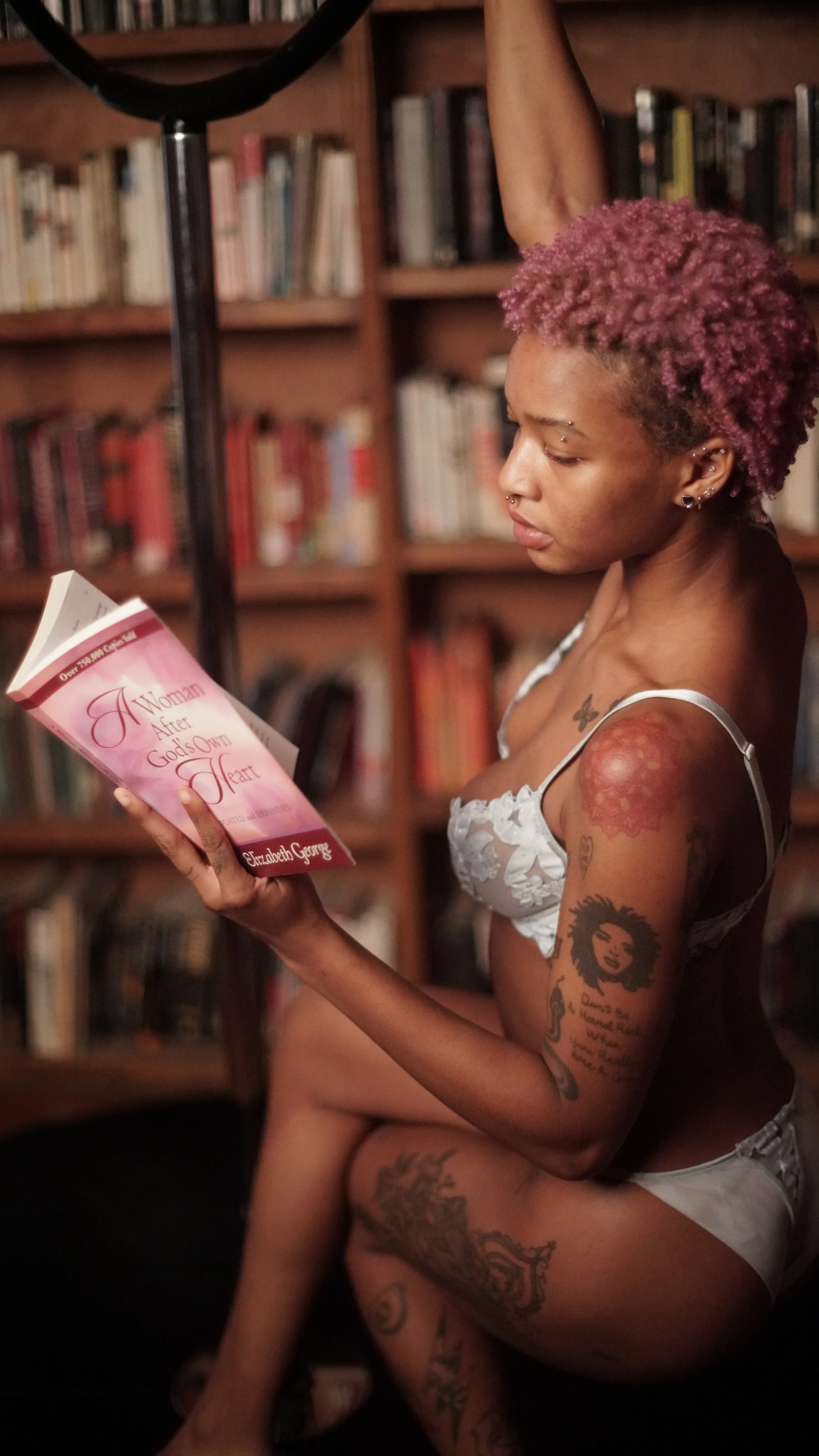 A woman with pink curly hair reading a book titled 'A Woman After God's Own Heart' in a room with bookshelves.