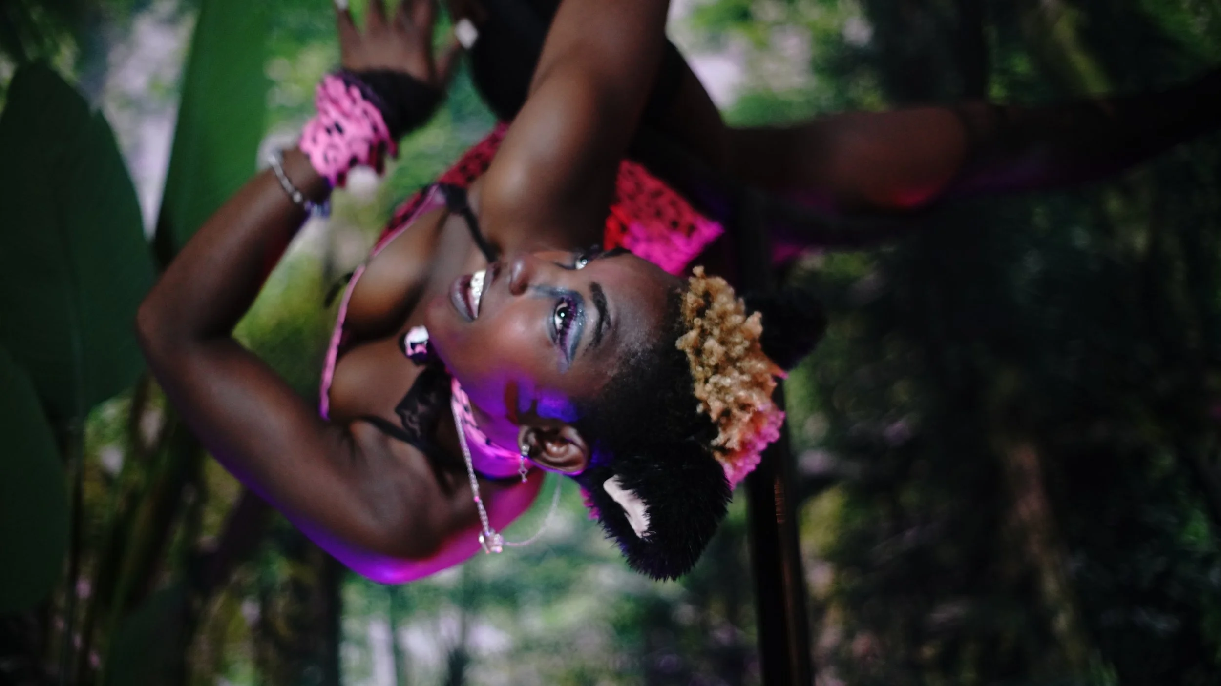 A woman with a unique hairstyle featuring pulled-up puffy sections and blonde highlights, dressed in a colorful outfit, lying on forest ground surrounded by green leaves and trees, smiling while looking up.