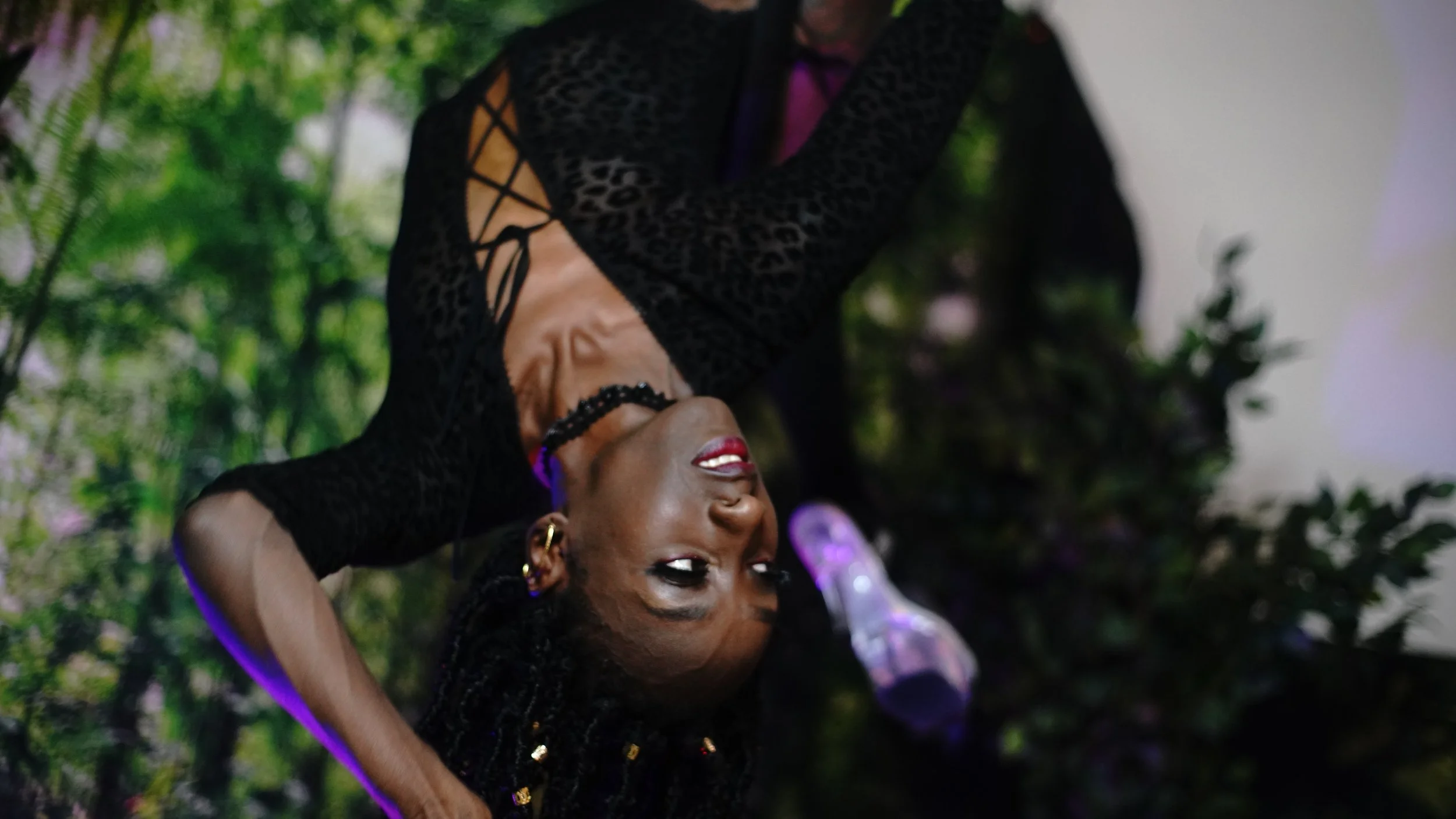 A woman with dark skin and braided hair adorned with gold beads, wearing a black lace dress with cut-out sleeves, is performing acro yoga upside down outdoors. She has red lipstick and is smiling with her eyes closed. The background features greenery