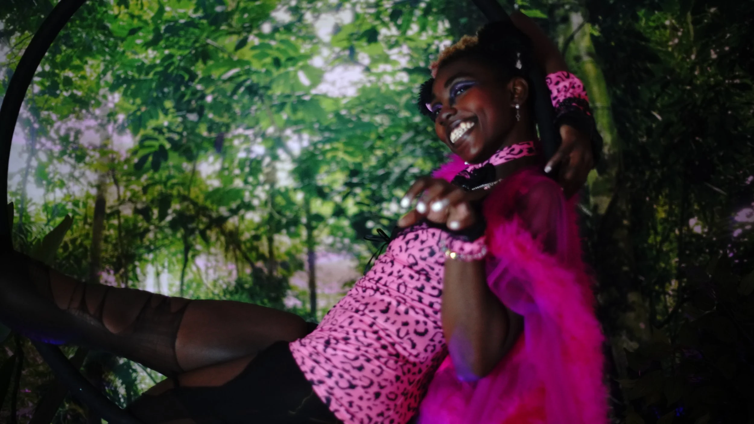 A woman in vibrant pink and leopard print clothing with makeup, jewelry, and hair accessories, smiling and posing in a lush green forest setting.