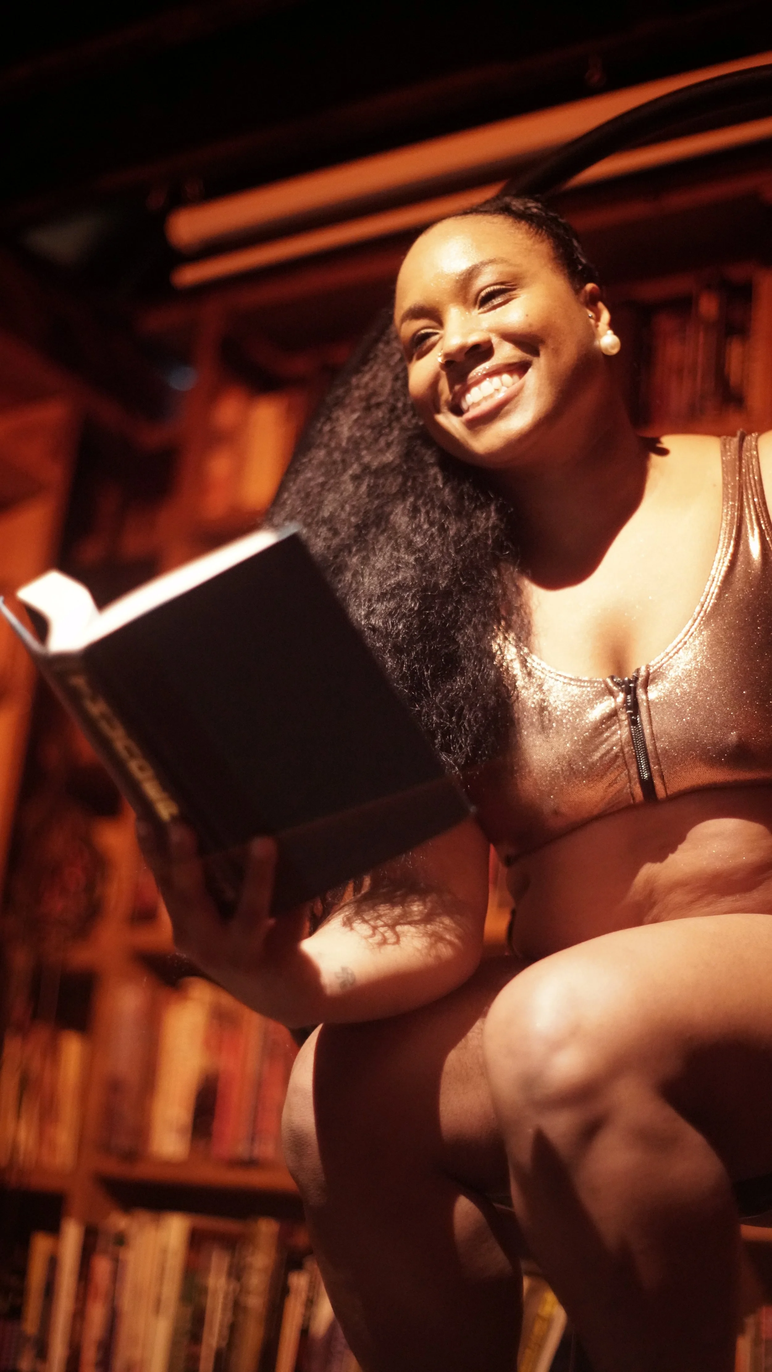 A smiling woman in a shiny, metallic top with a zipper, sitting in a room with wooden bookshelves filled with books, holding an open book in her hand.