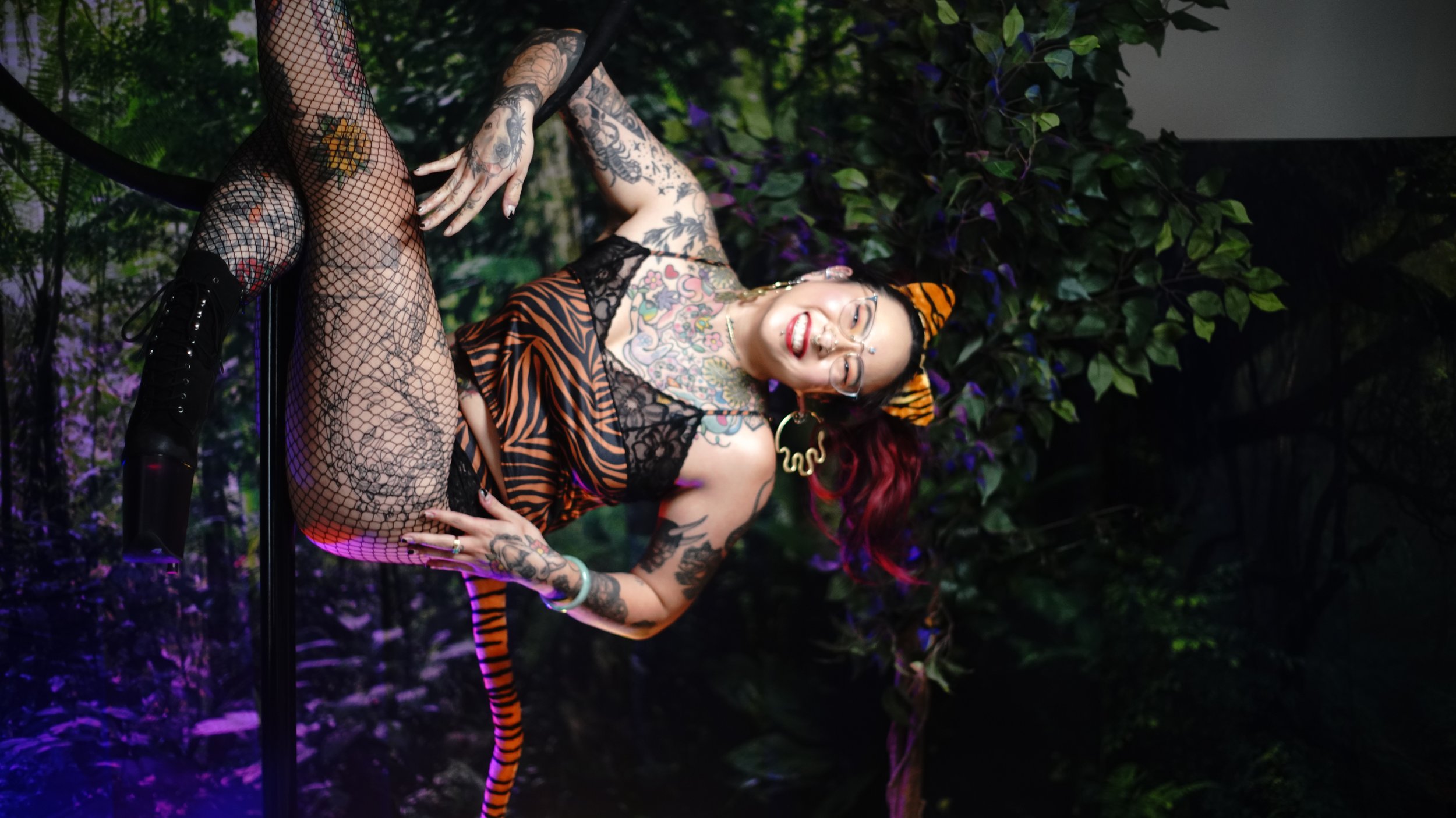 A woman with tattoos wearing tiger-striped costume, fishnet stockings, black boots, and tiger ears headband, smiling and sitting on a black pole against a jungle background with flowers and greenery, with purple lighting.