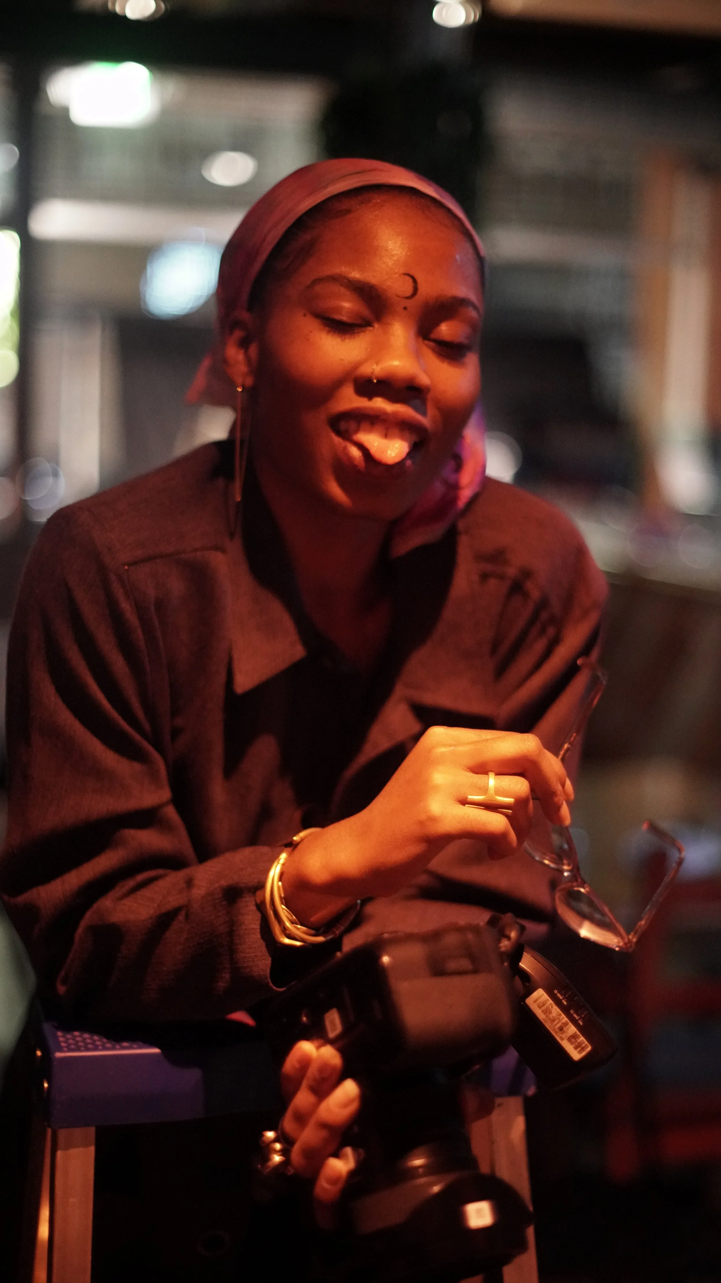 Woman with short hair wearing a headscarf, smiling playfully with tongue out, holding eyeglasses and a camera in a low-light indoor setting.