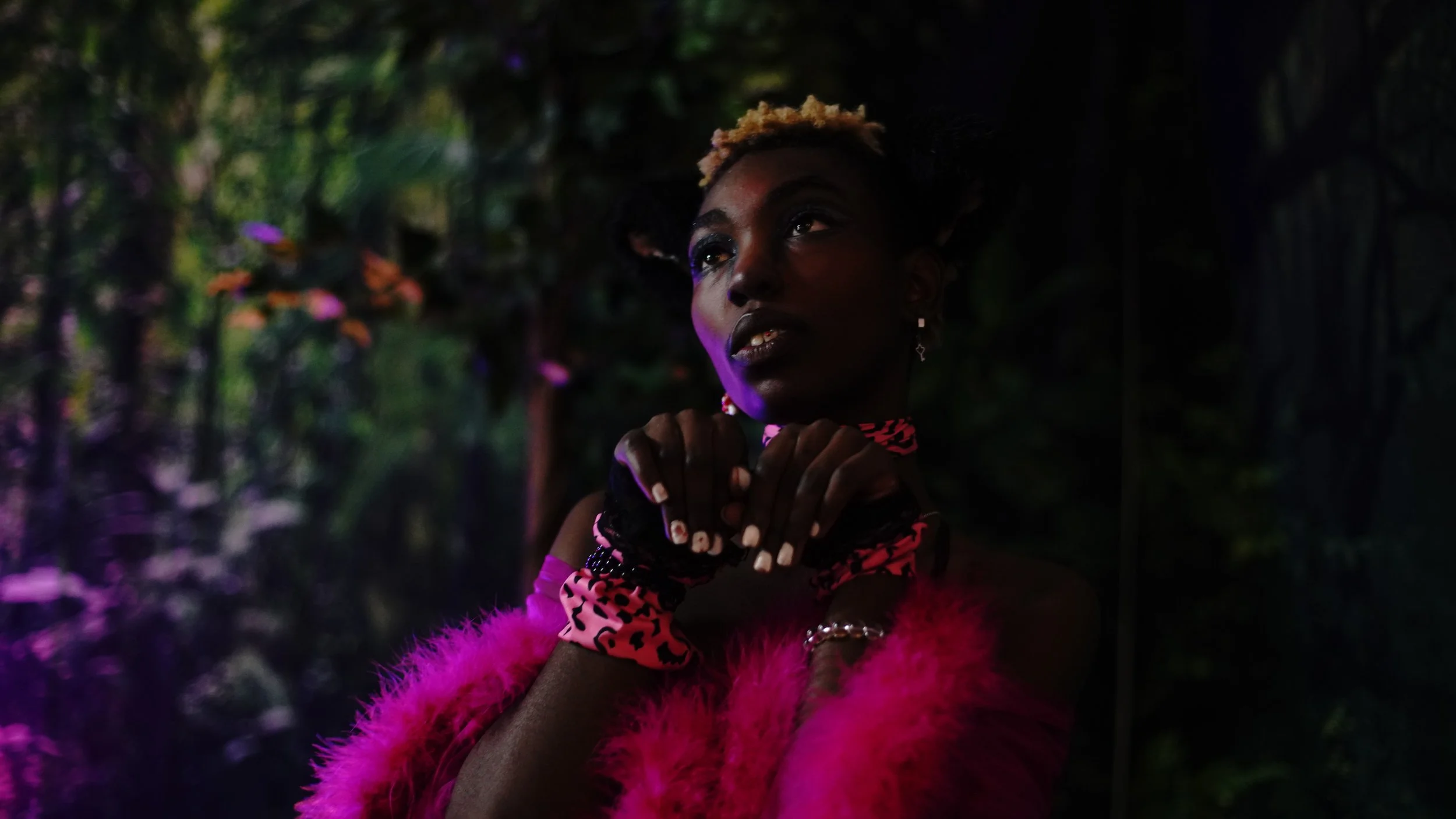 A woman with dark skin in a pink furry coat, black gloves, and leopard print accessories, posing with her hands near her face in a dark, lush garden background with purple lighting.