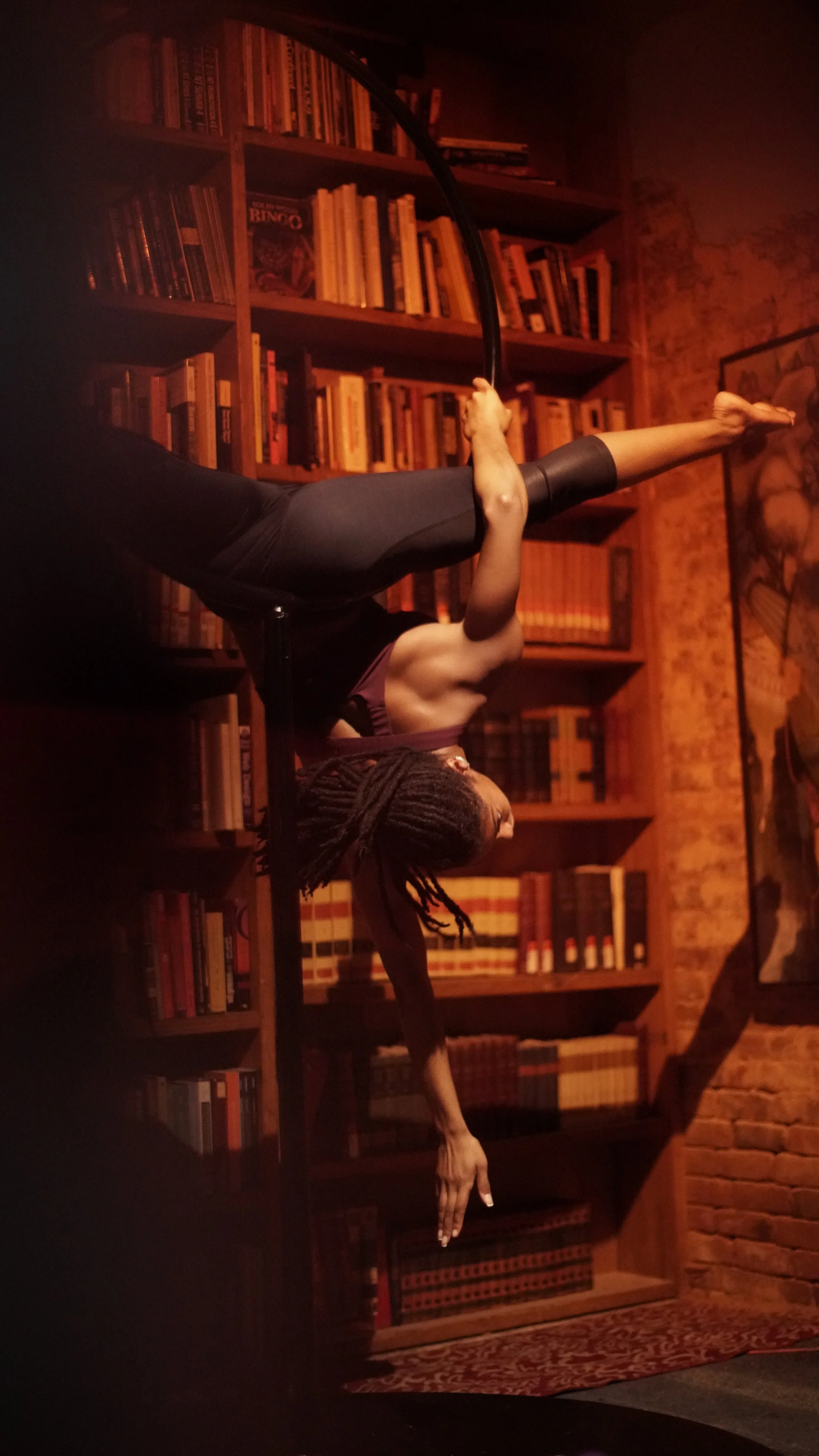 A woman with dreadlocks performing an aerial pose on a hoop in a cozy, dimly lit room with wooden bookshelves filled with books and a brick wall.