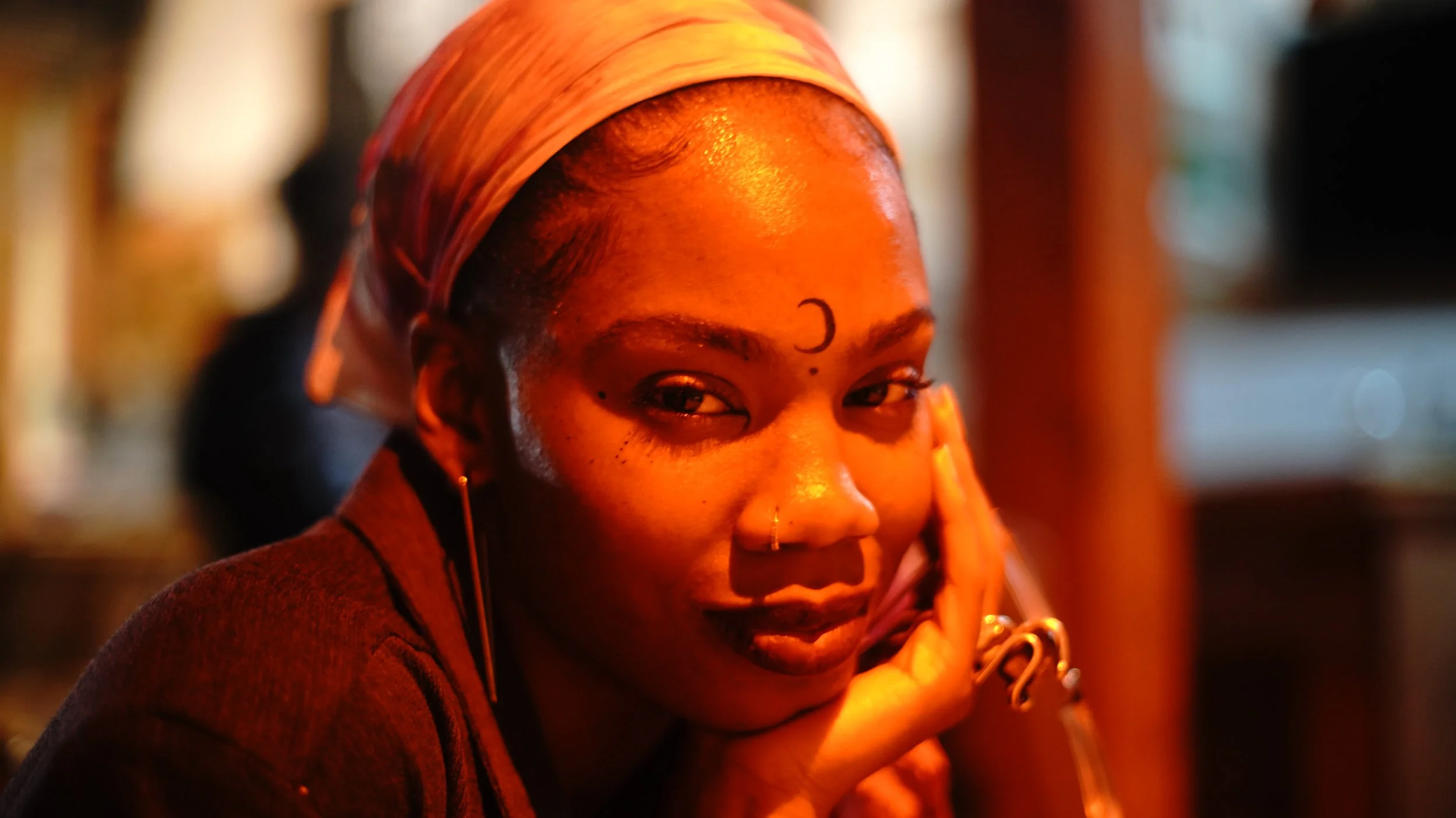 Close-up of a woman with dark skin, wearing a colorful headscarf, looking at the camera with a slight smile, resting her chin on her hand, in a warm, softly lit setting.