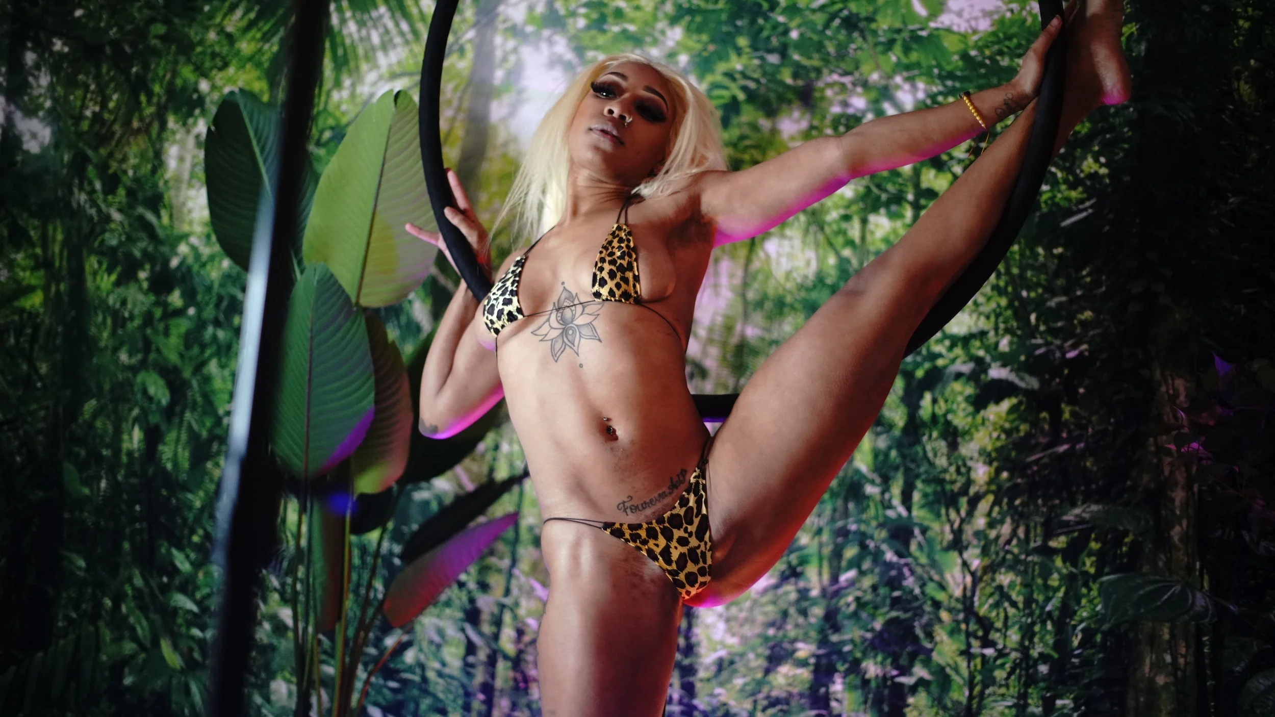 A woman with blonde hair in a leopard print bikini performing a high leg stretch in front of a jungle backdrop. She has tattoos and jewelry, and is holding a black hoop.