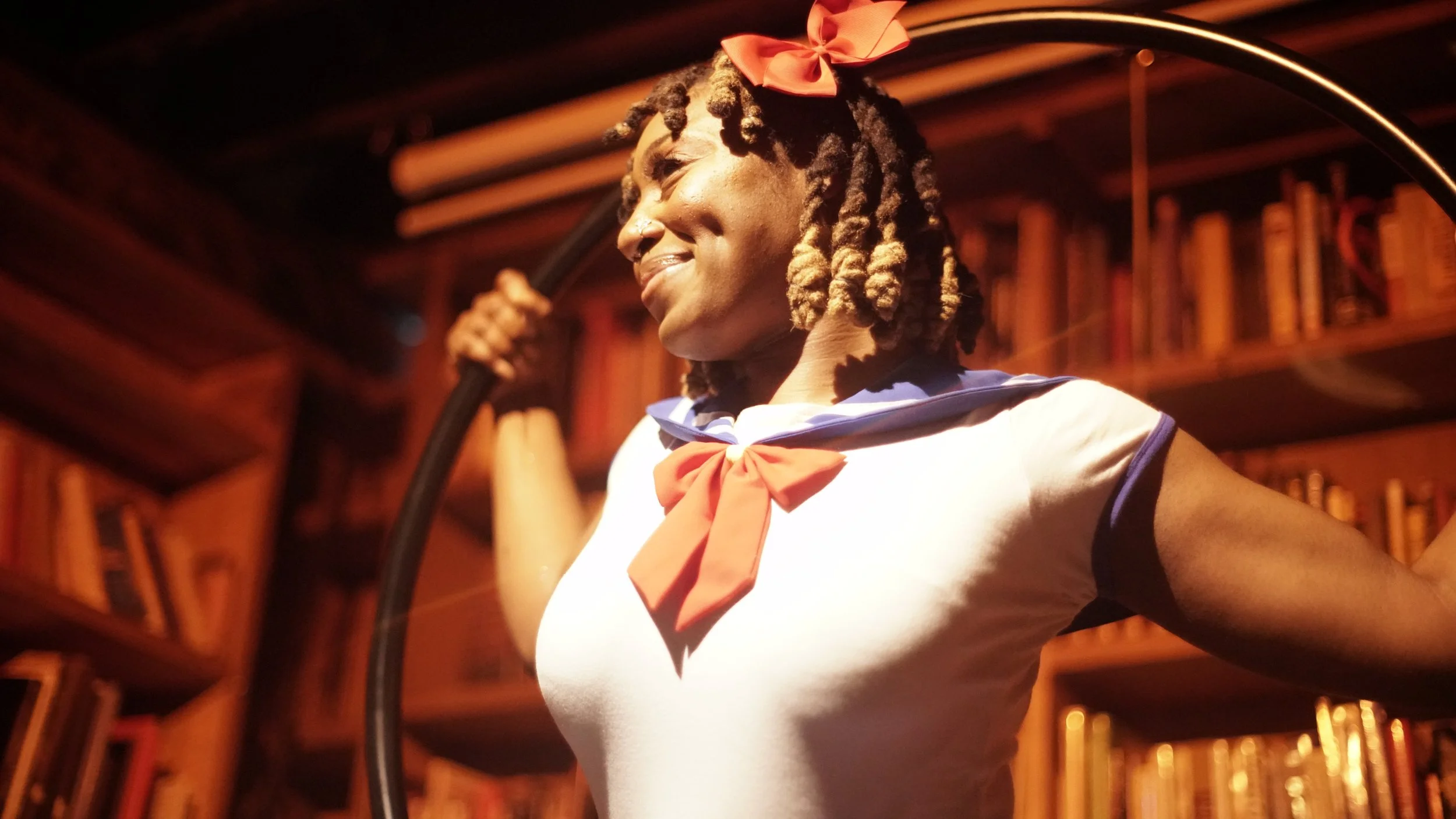 A person with dreadlocks wearing a red bow and a sailor collar shirt, standing in a warm-colored library, smiling and holding a hoop.