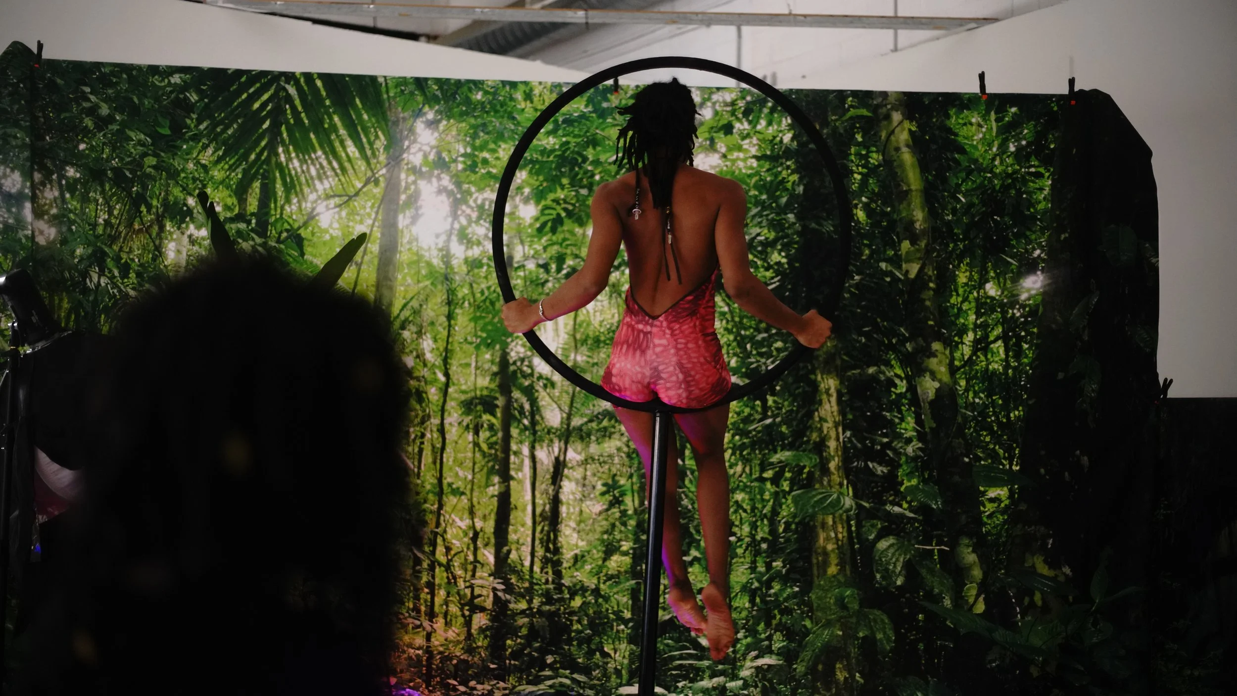 Person with long dreadlocks wearing a backless red outfit performing aerial silks against a jungle-themed backdrop.