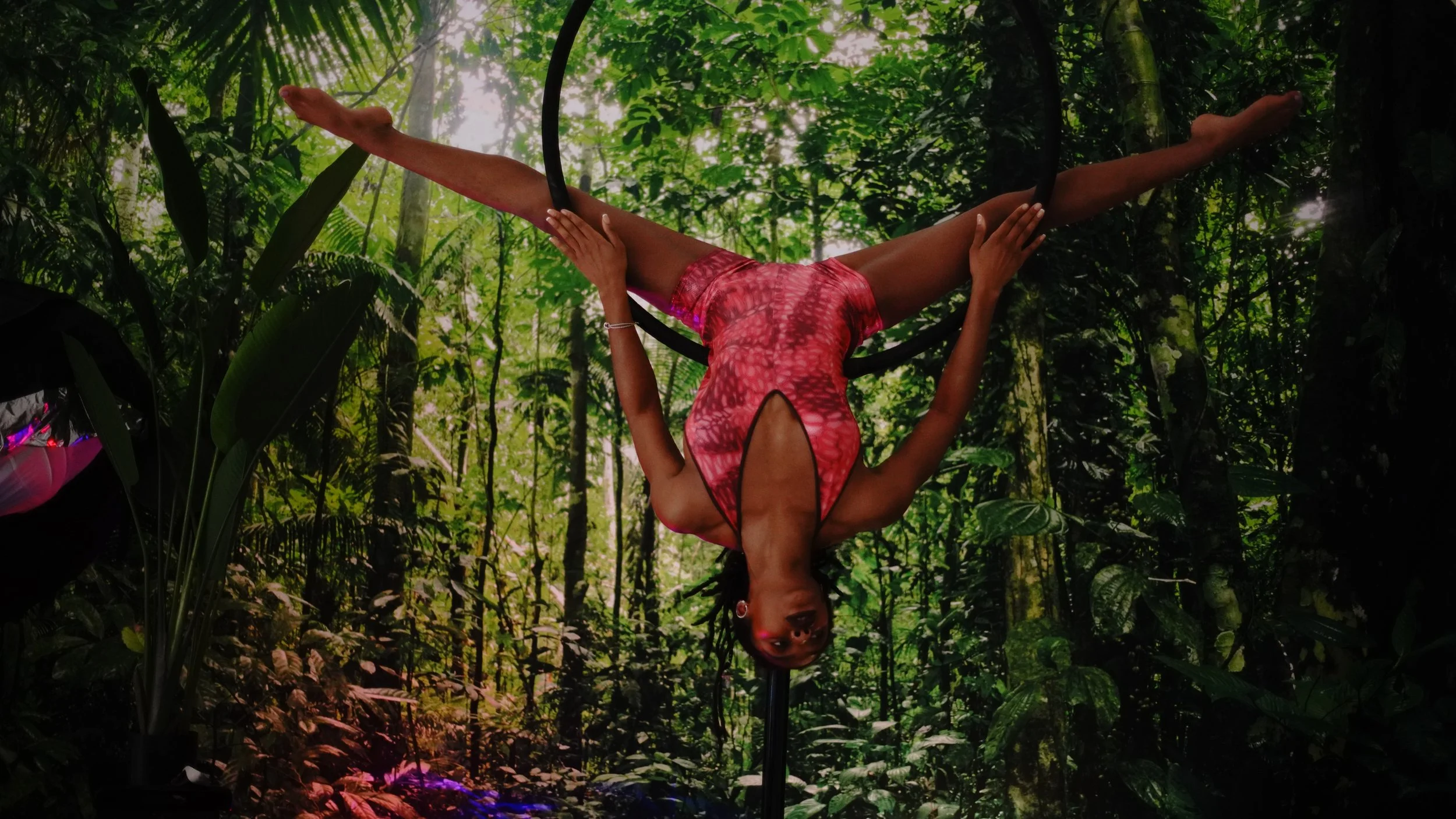 A woman dressed in a red patterned sleeveless top and pink shorts practicing aerial yoga on a black hoop suspended in a dense jungle setting.