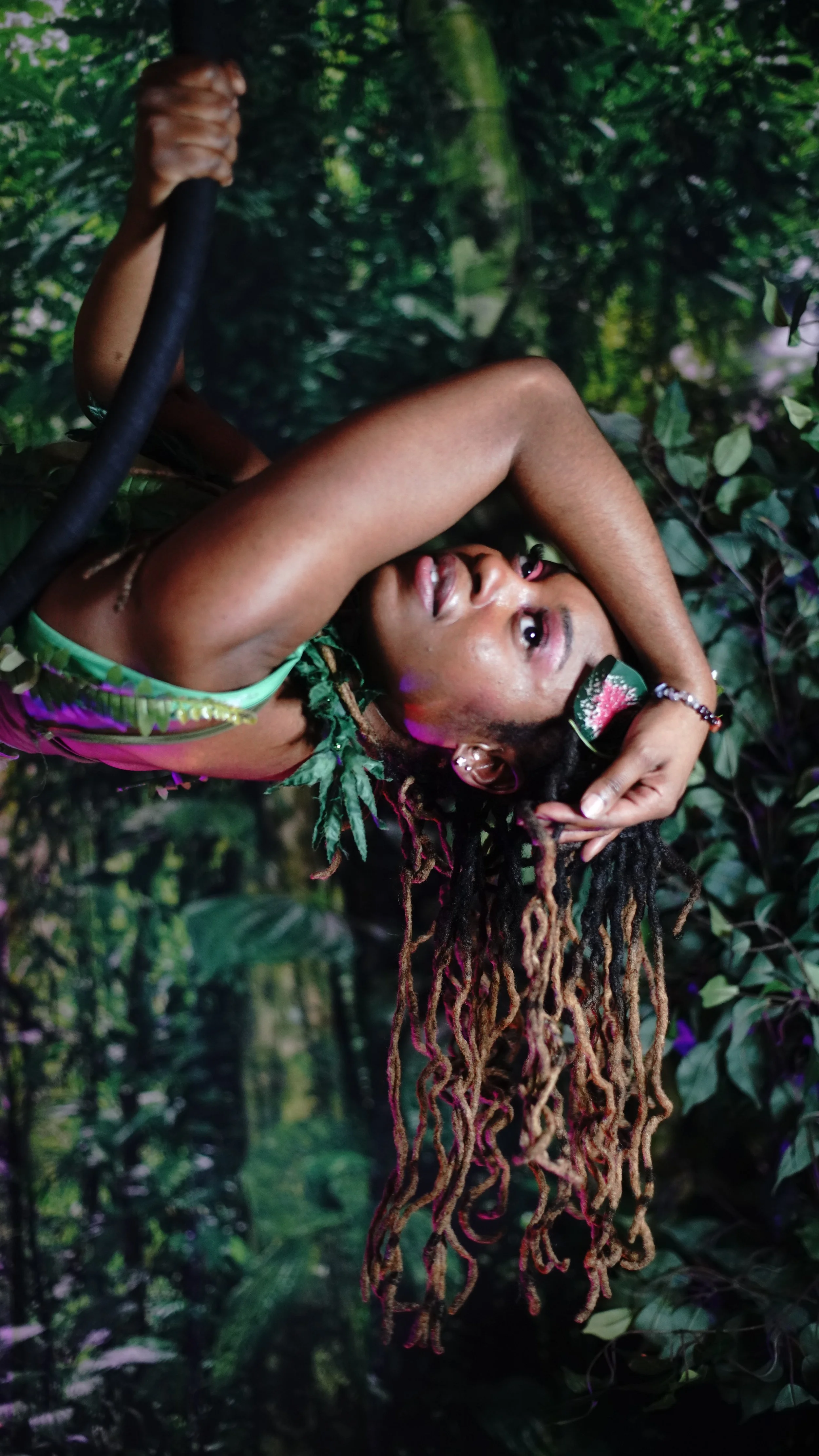 A woman with long dreadlocks in a lush jungle setting, holding a small axe and smiling.