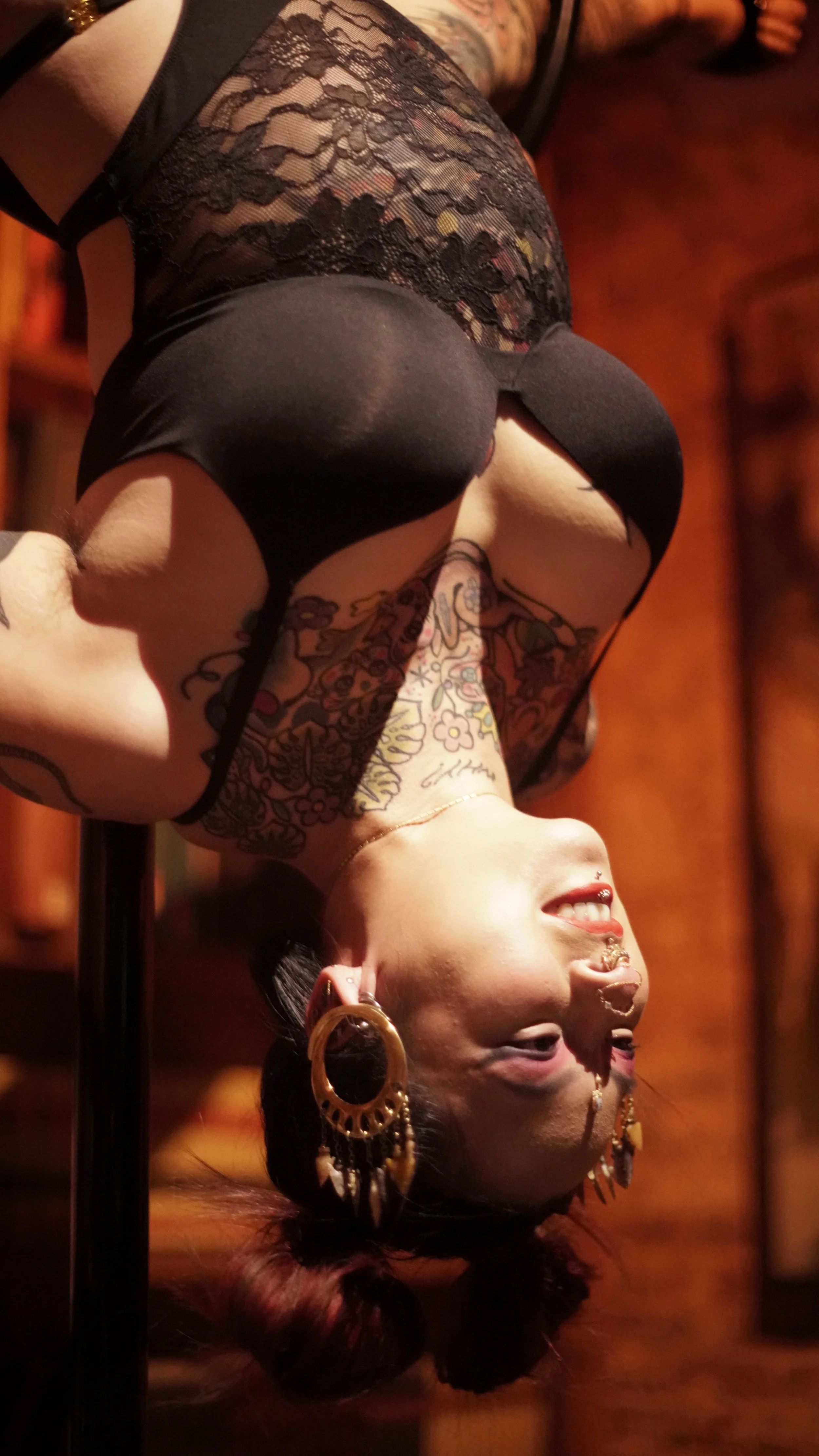 A woman with tattoos and large hoop earrings is performing a pole dance upside down, smiling in a dimly lit room with wooden walls.