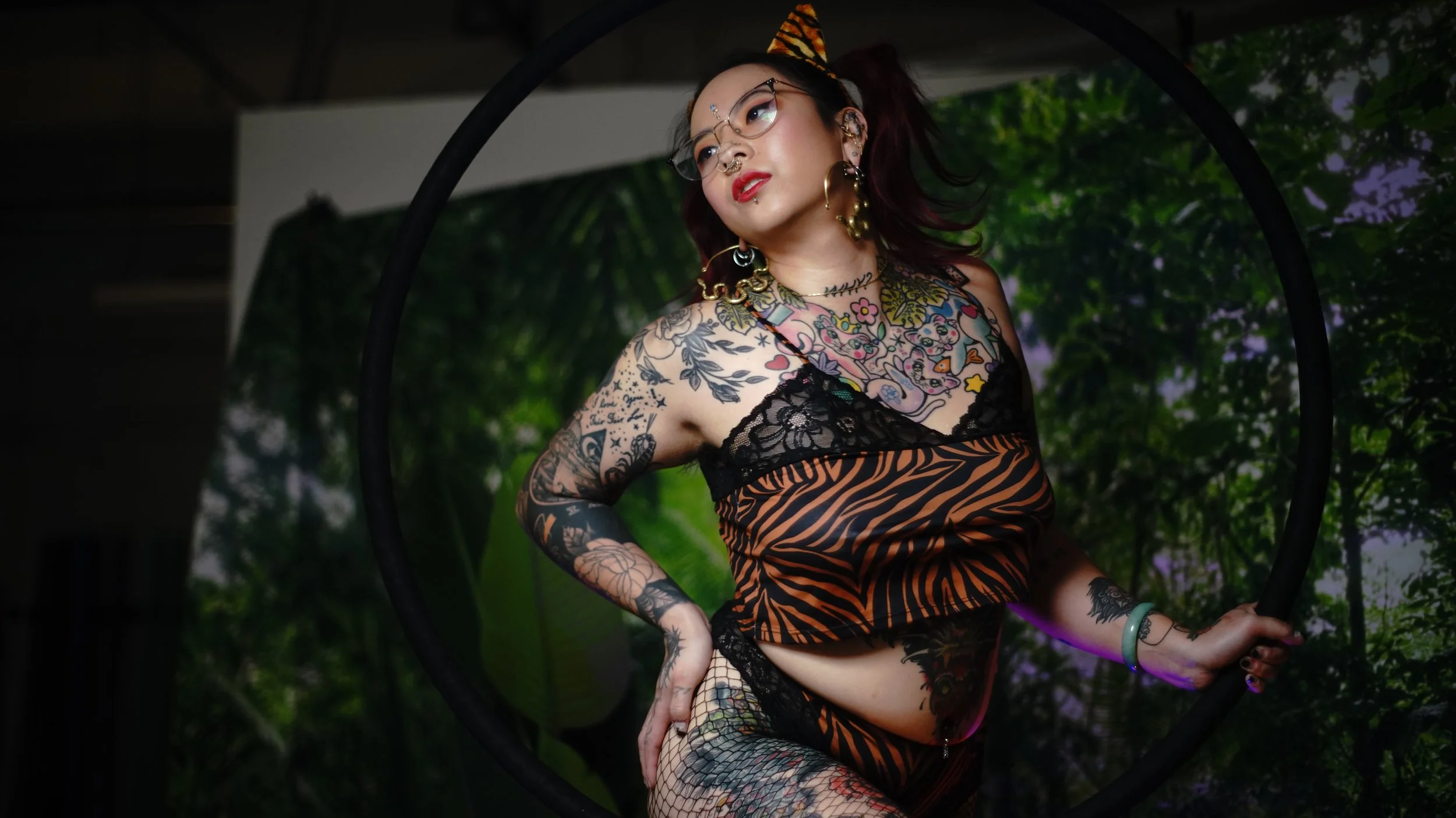 A tattooed woman with glasses, lace and animal print clothing, posing through a black circular frame, with a background of lush green tropical plants.