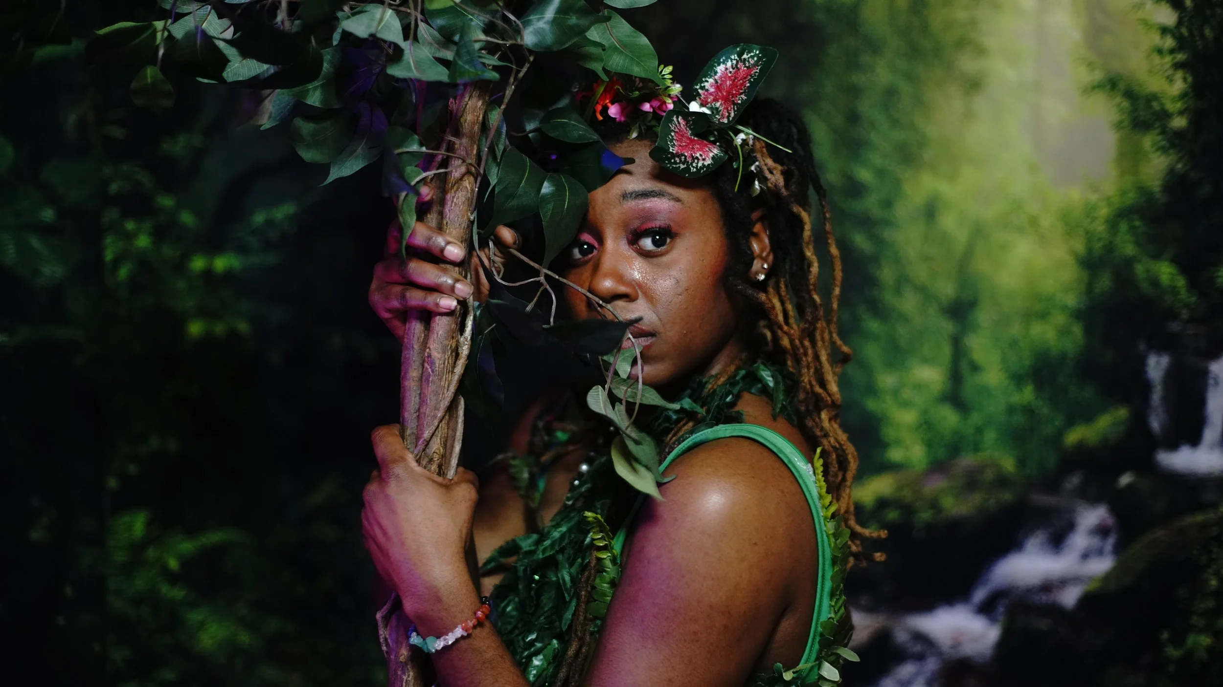 A woman with dreadlocks and a green dress partially hidden behind a tree branch with leaves, in a lush forest with a creek in the background.