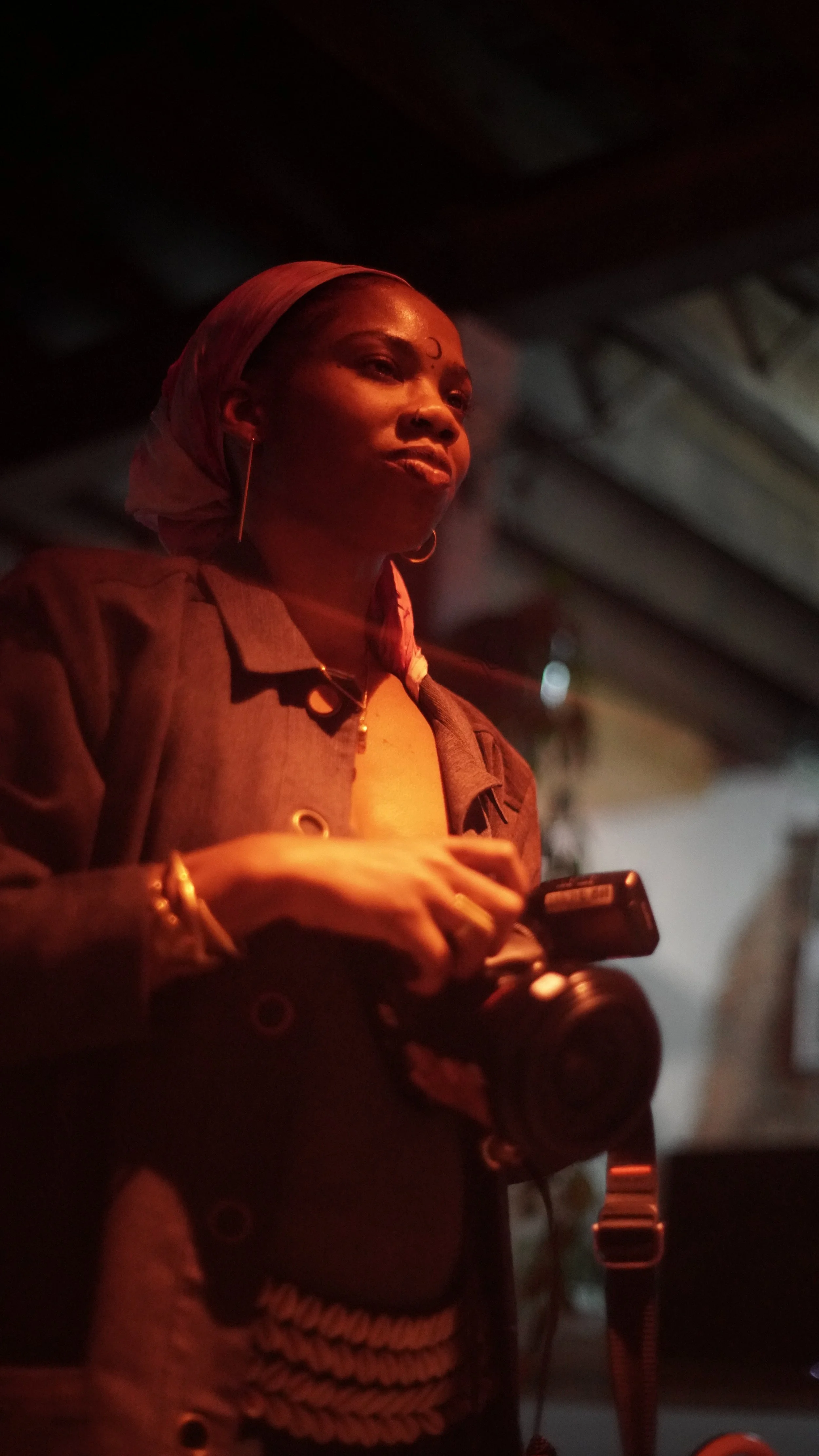 A woman wearing a headscarf and hoop earrings, holding a camera and a phone, standing in a dimly lit space.
