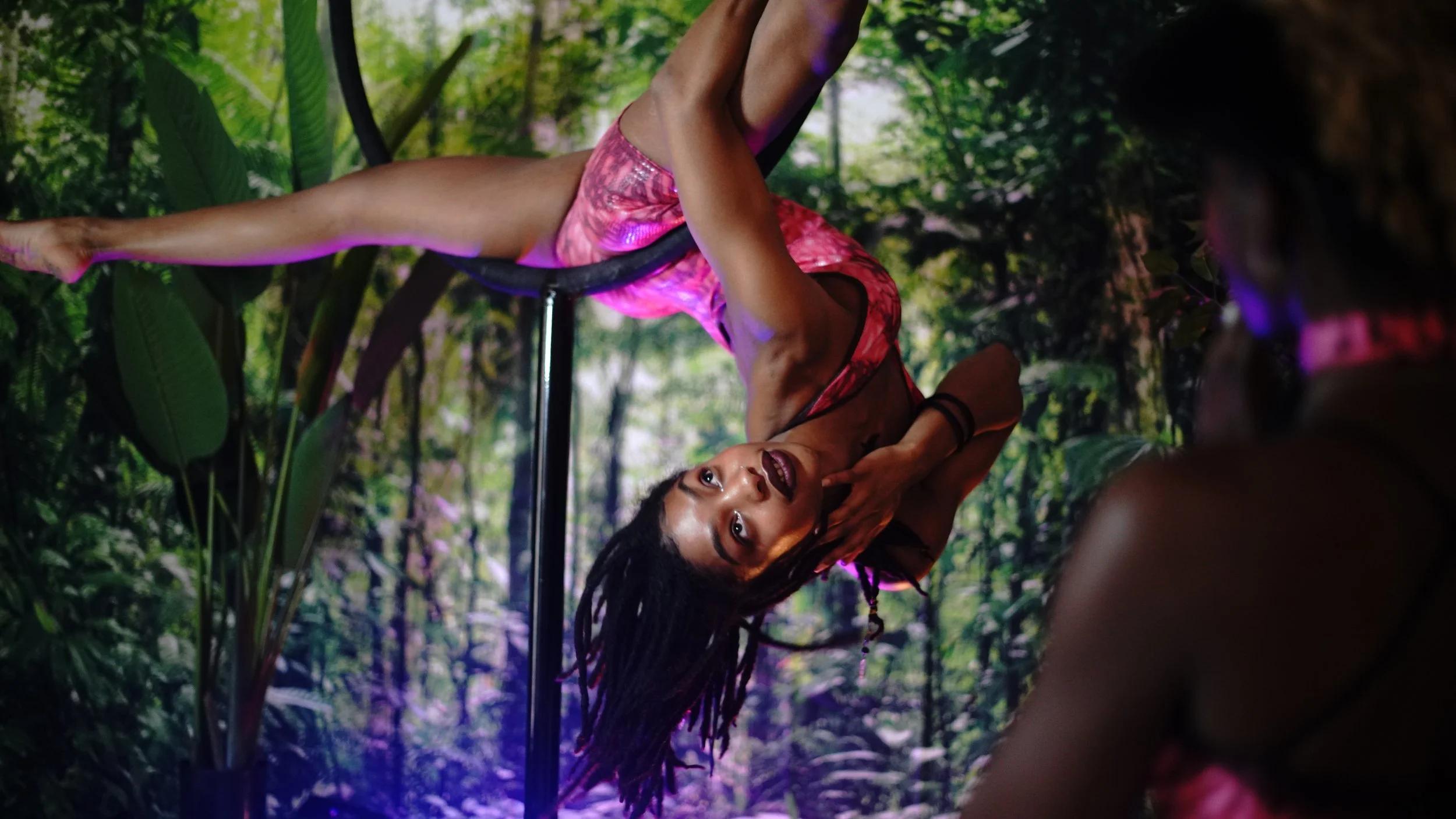 A woman with dreadlocks practicing pole dance in a lush jungle setting during a performance, with a person in the foreground watching.