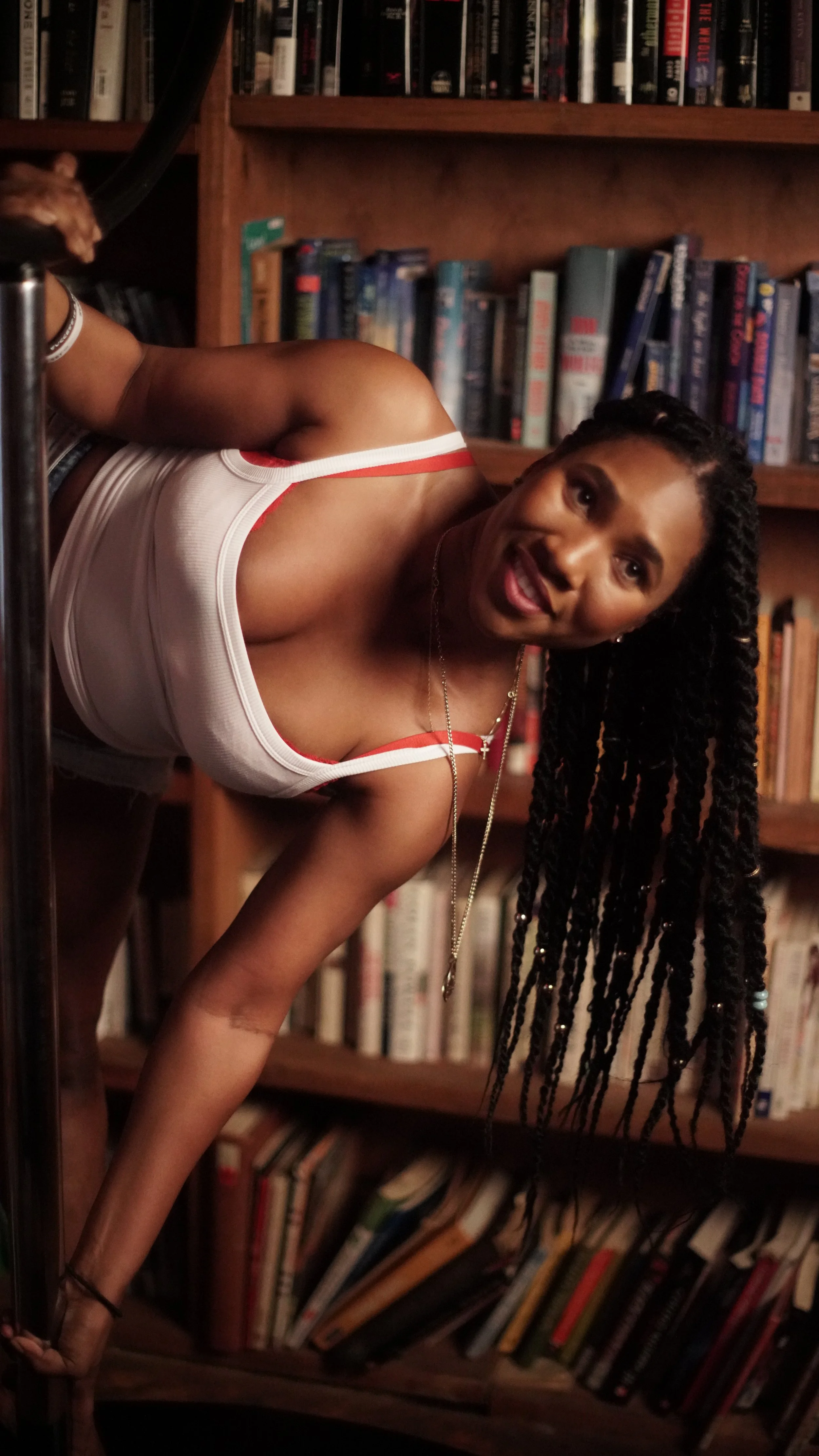 A woman with long, braided hair leaning forward and smiling in front of a bookshelf filled with books.