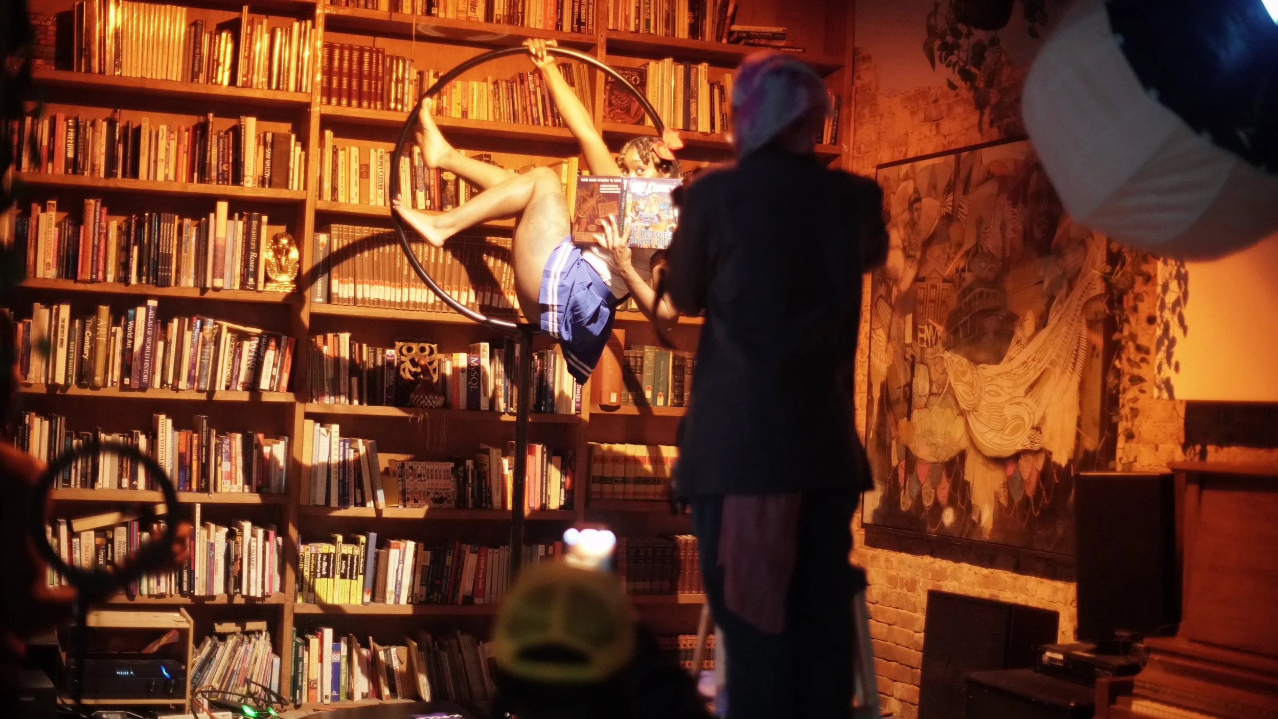 A woman inside a bookshop reads a book while sitting inside a circular metal art installation, with the backdrop of a large bookshelf filled with books. A person wearing dark clothing and glasses stands nearby, partially reflecting the scene. The set