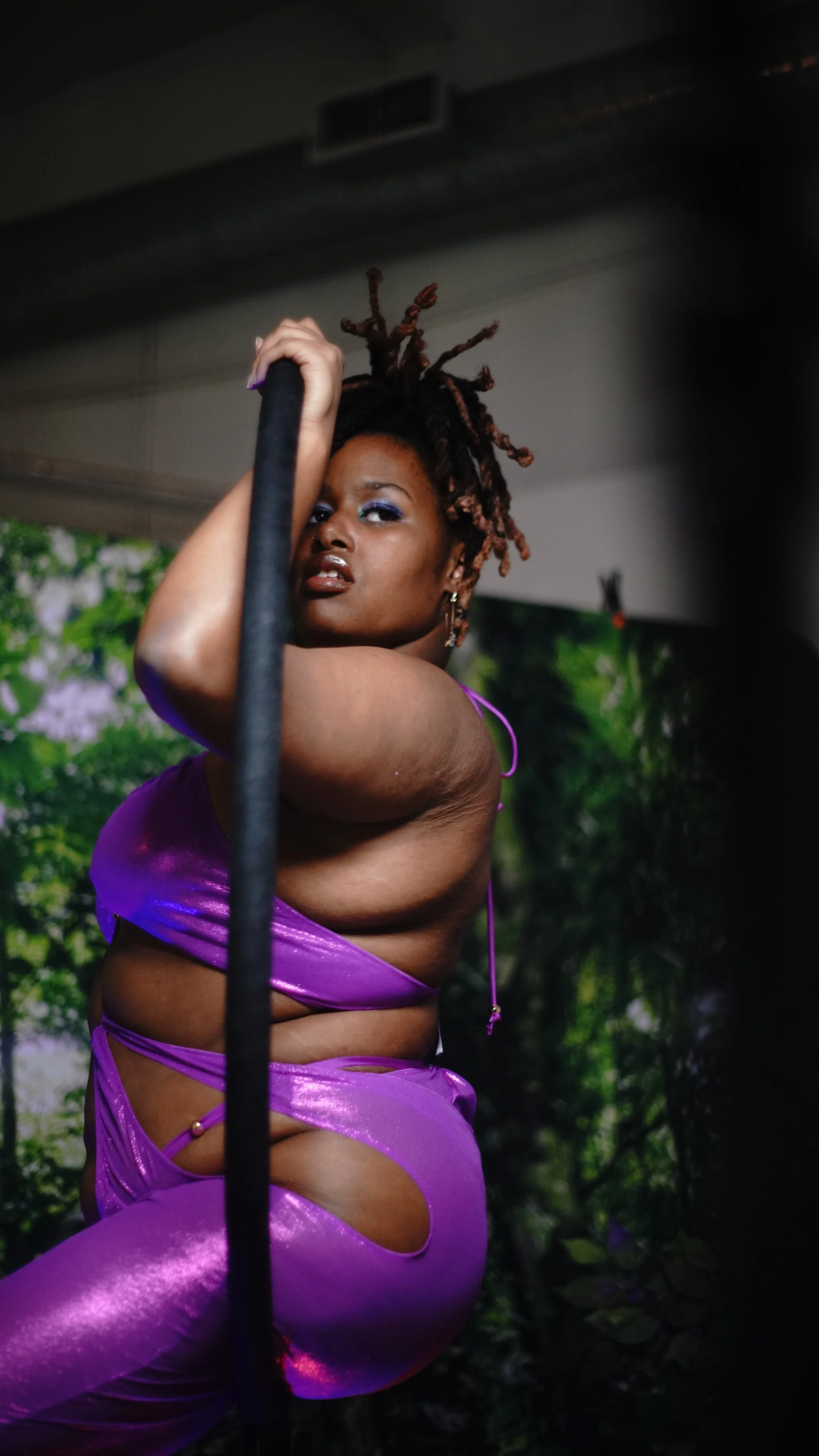 A woman with dreadlocks hanging over her shoulder, wearing a shiny purple outfit, holding onto a black pole against a green nature-themed background.