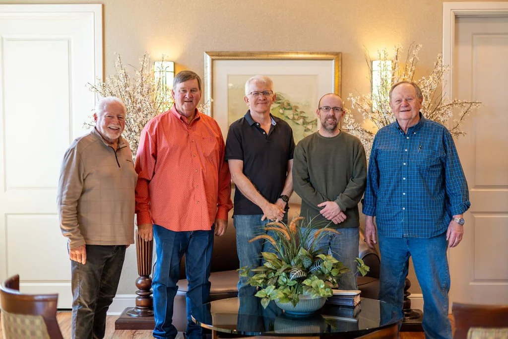 Five men standing in a living room with a floral arrangement on a glass table in front of them, behind them are wall-mounted lights, a framed picture, and decorative plants.