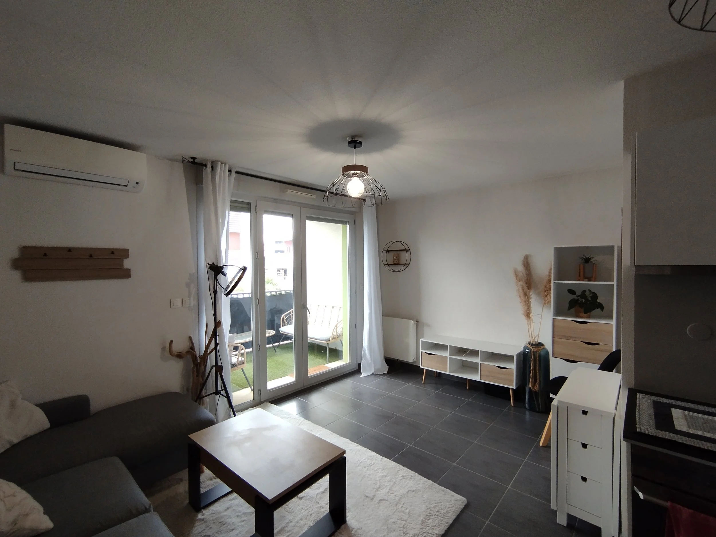 Salon avec canapé gris, table basse, livres, étagère blanche et porte-fenêtre laissant entrer la lumière naturelle.