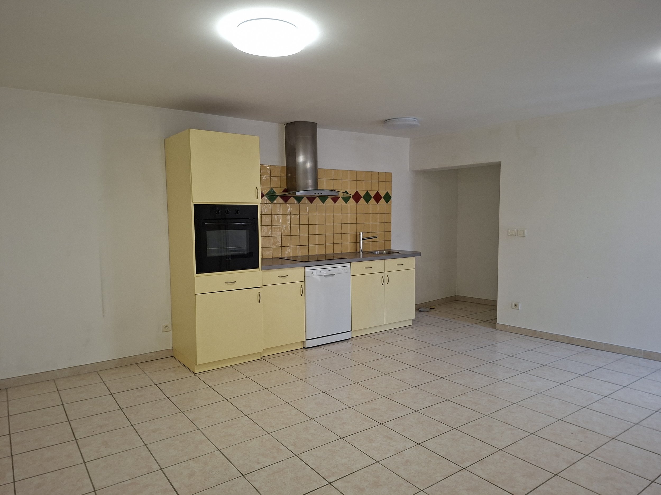 Cuisine simple avec des armoires jaunes, un four encastré, un évier, une hotte, un dosseret en carreaux beige avec une bande de carreaux verts et rouges, et un espace_vide.