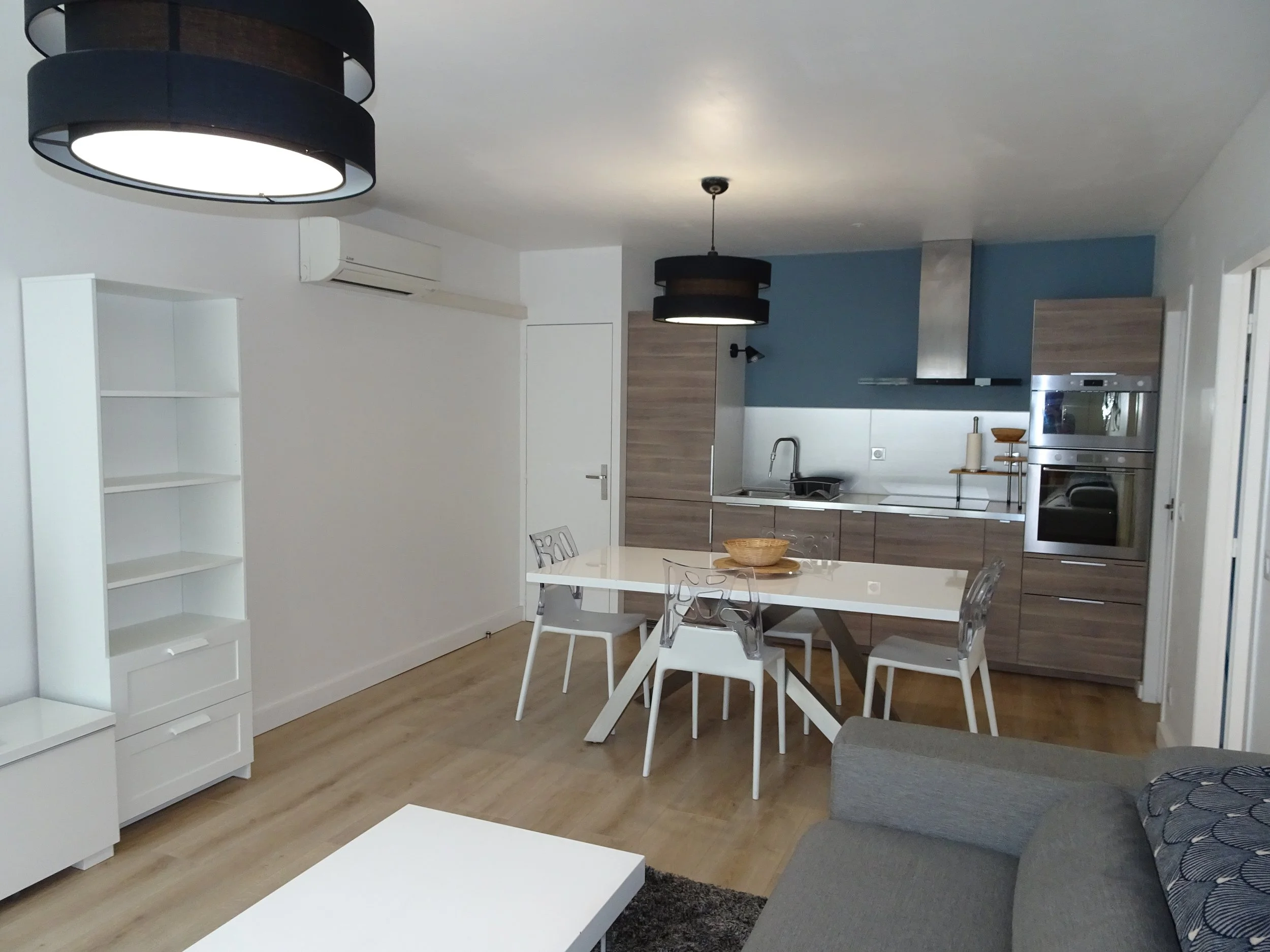 Un intérieur de cuisine moderne avec des meubles en bois clair, un plan de travail blanc, une table blanche entourée de chaises transparentes, et un coin salon avec un canapé gris. Il y a deux plafonniers noirs suspendus au plafond, un réfrigérateur encastré, un four, un micro-ondes, et une partie du mur peint en bleu.