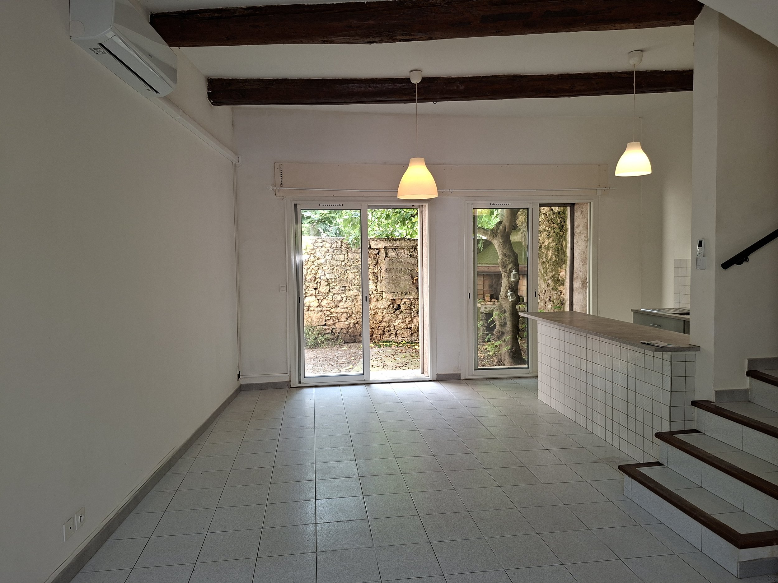 Une pièce intérieure lumineuse avec des fenêtres donnant sur un jardin. Il y a deux lampes suspendues, un mur blanc, un sol en carrelage clair, une cuisine partielle à droite, un escalier en bois à droite.