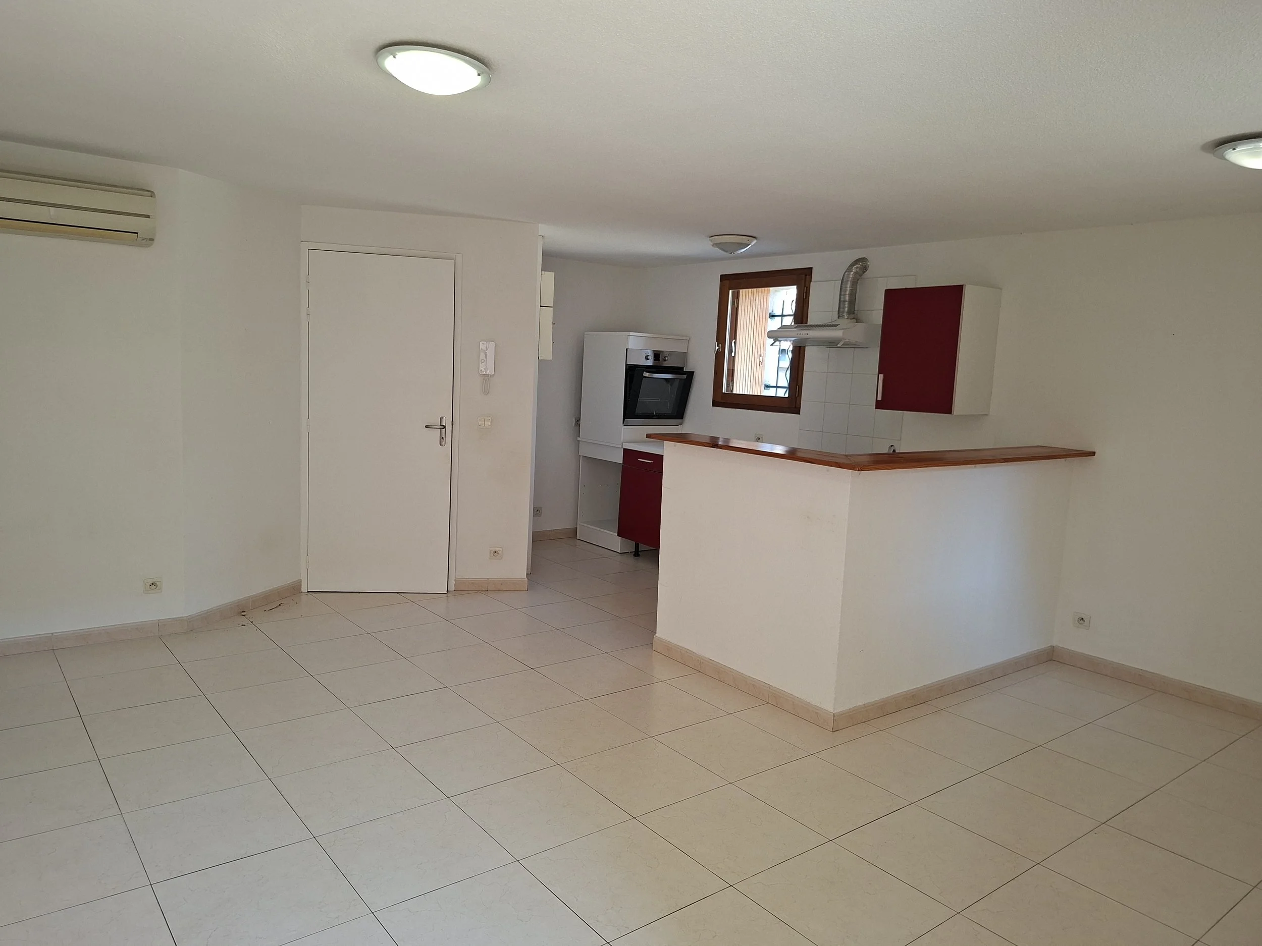 Intérieur d'une pièce de cuisine avec murs blancs, sol en carreaux beige, un ventilateur de plafond, une fenêtre en bois, une hotte de cuisine, un four encastré, un comptoir en bois, et une porte blanche.