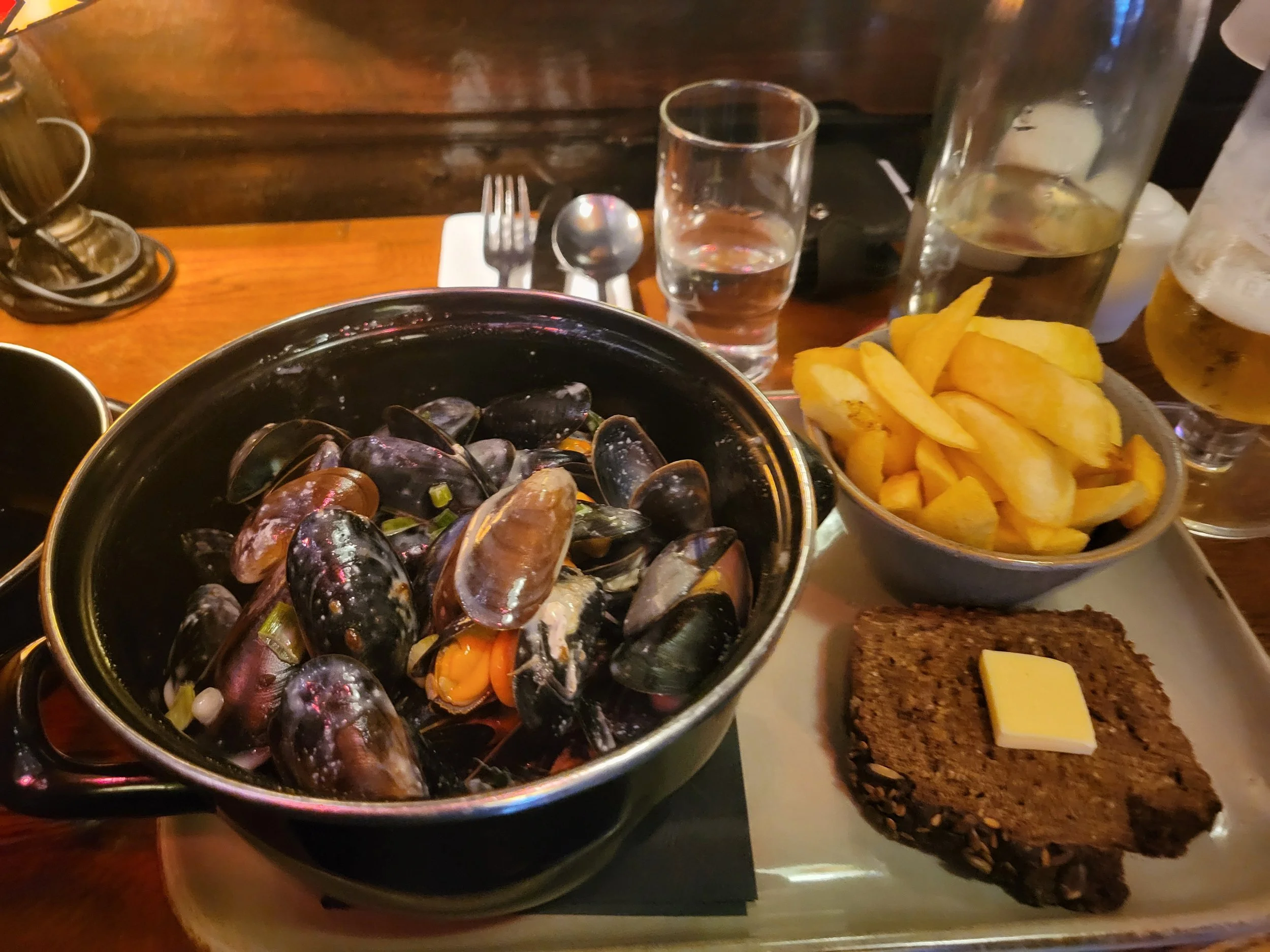 A plate of cooked mussels in a black dish with French fries, a slice of bread with butter, and a small notepad with silverware and glasses on a wooden table.