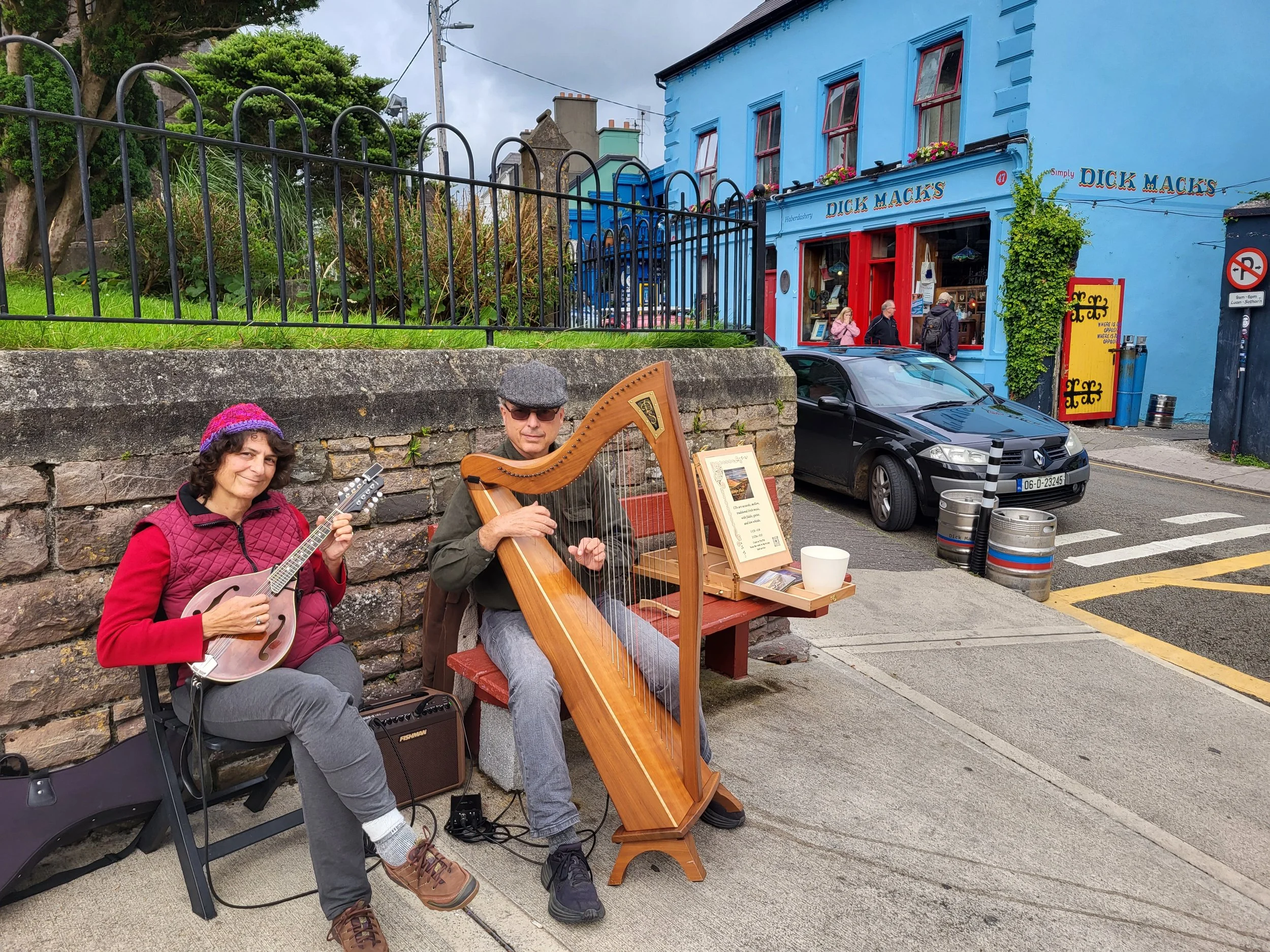 Two street musicians sit on a sidewalk in front of a stone wall, playing instruments. The woman on the left holds a guitar, while the man on the right plays a harp. Behind them is a colorful blue building with a shop named 'Duck Macks' and pedestrians walking by.