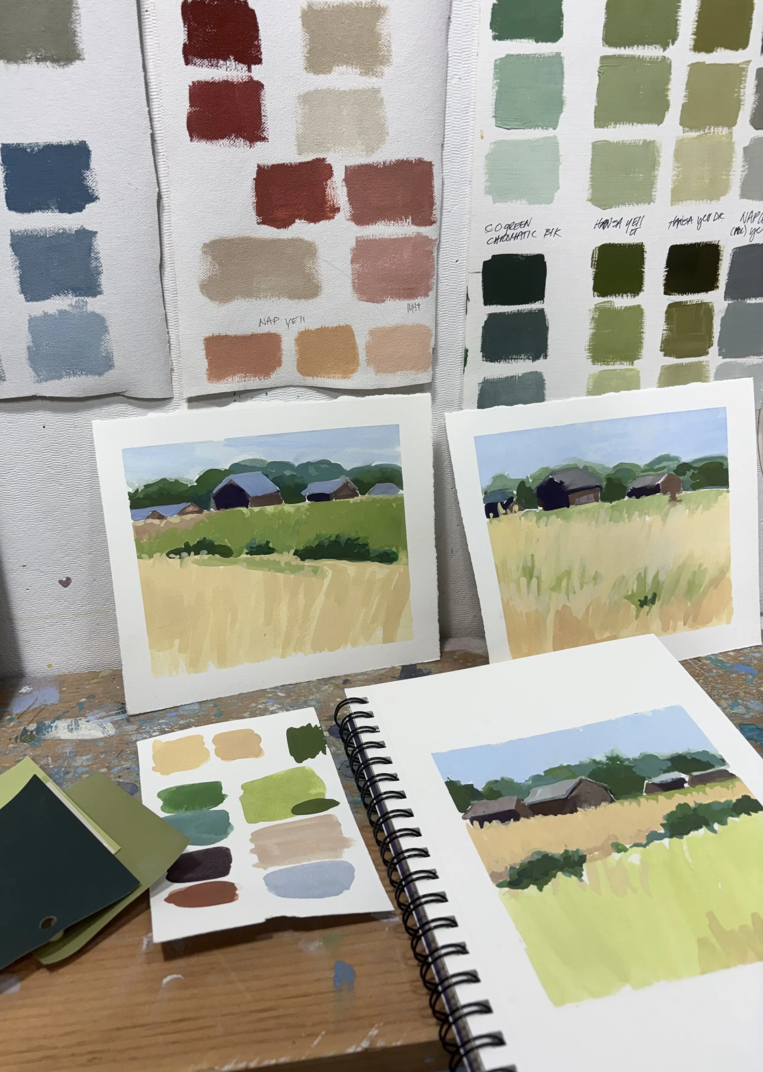 Paintings and color swatches of rural landscape scenes with barns and grass, made with acrylic or oil paints, displayed on a wall and table.