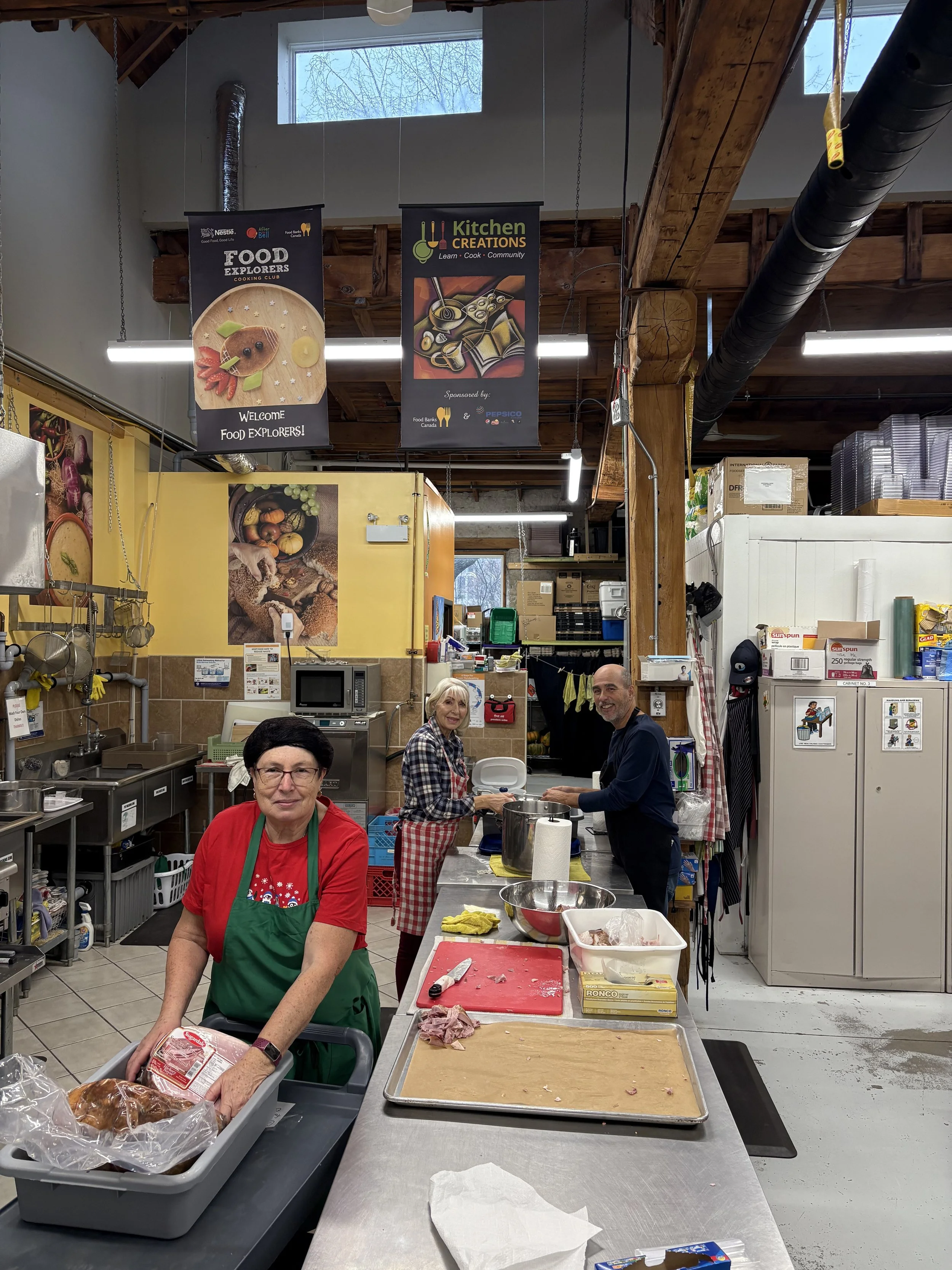 Three people in a kitchen volunteering for Food Explorers. The woman in the foreground wears a red shirt and green apron, holding a loaf of bread in a plastic bag. The woman in the middle, with gray hair, wears a plaid shirt and red checkered apron, standing beside a man with a bald head and gray beard. They are preparing food at a counter with various kitchen tools.