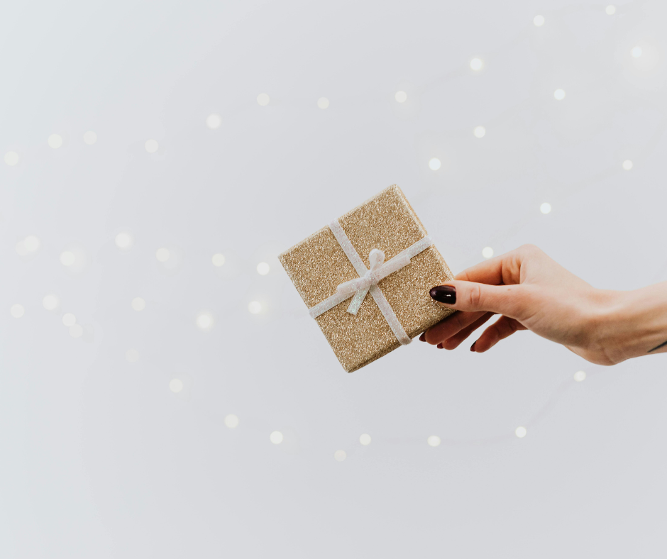 A hand holding a small, gold, glittery gift box with a white ribbon against a blurred background with white lights.