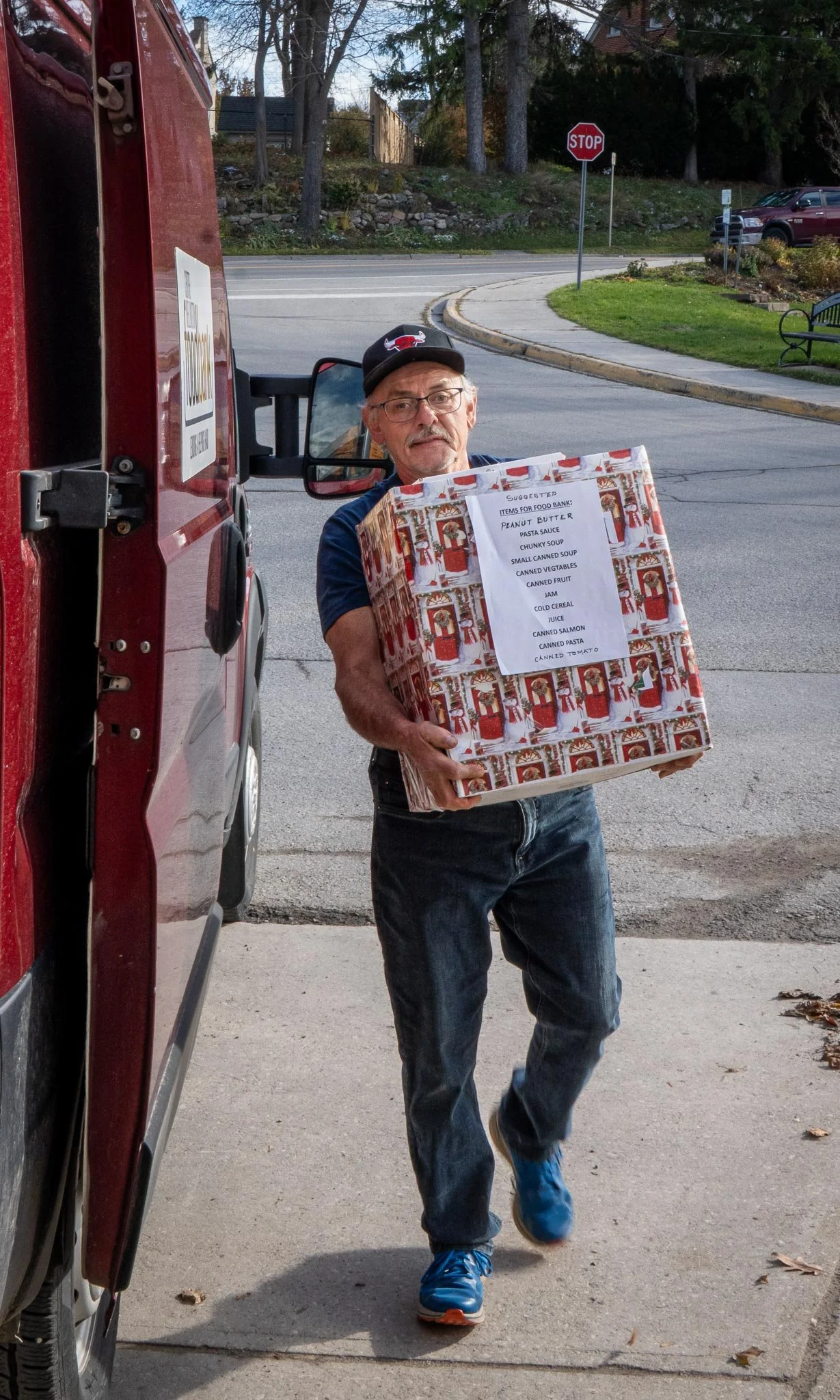 Man wearing glasses and a baseball cap carrying a large box with a Christmas-themed design. The box has a list of food items attached to it, indicating it is for a food bank donation.
