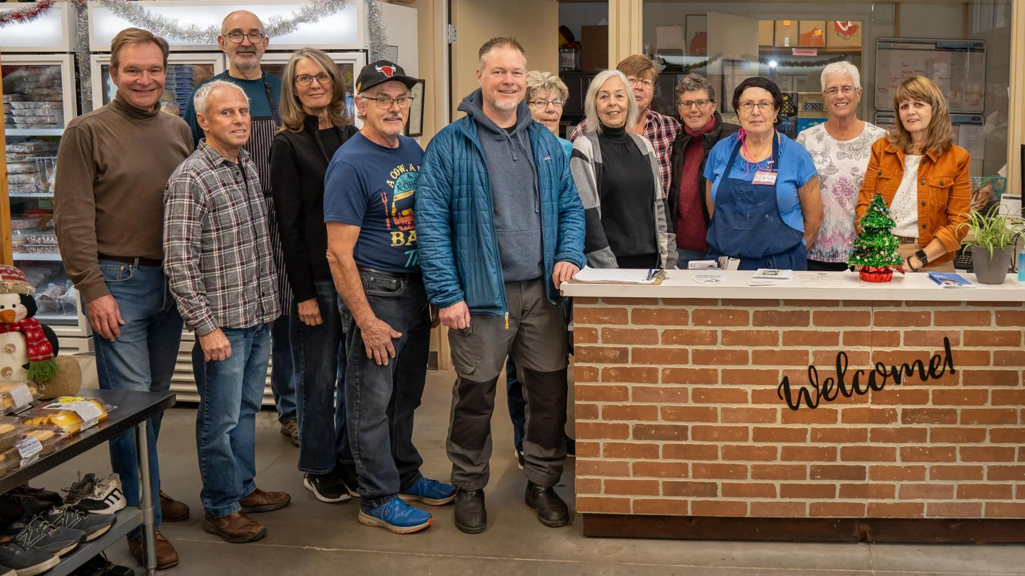 Group of people standing in line behind a brick counter with a 'Welcome' sign, some wearing aprons, in a community or retail setting with Christmas decorations.