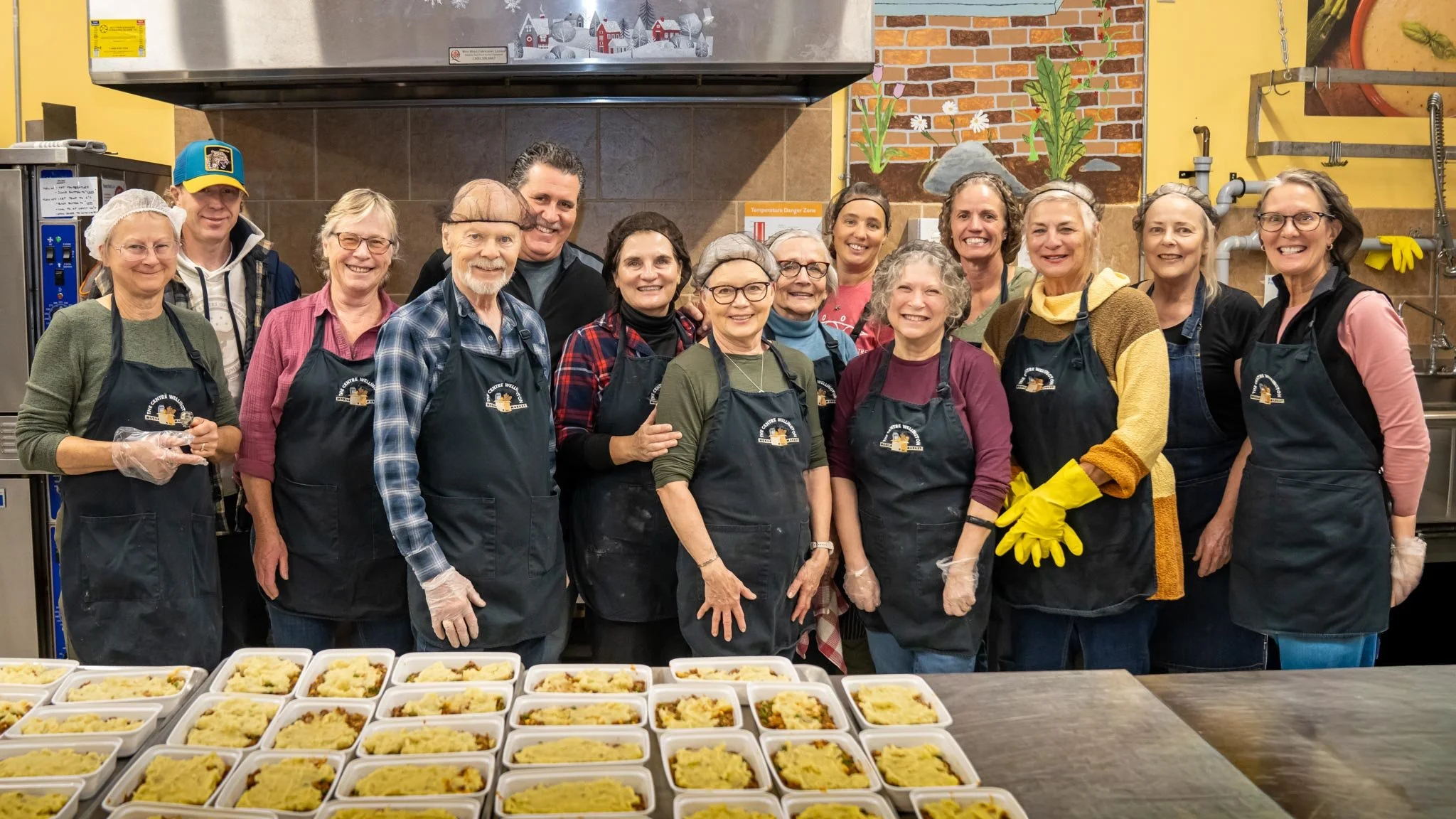 Group of volunteers smiling in a kitchen, wearing aprons and gloves, with prepared food containers on the table in front of them.