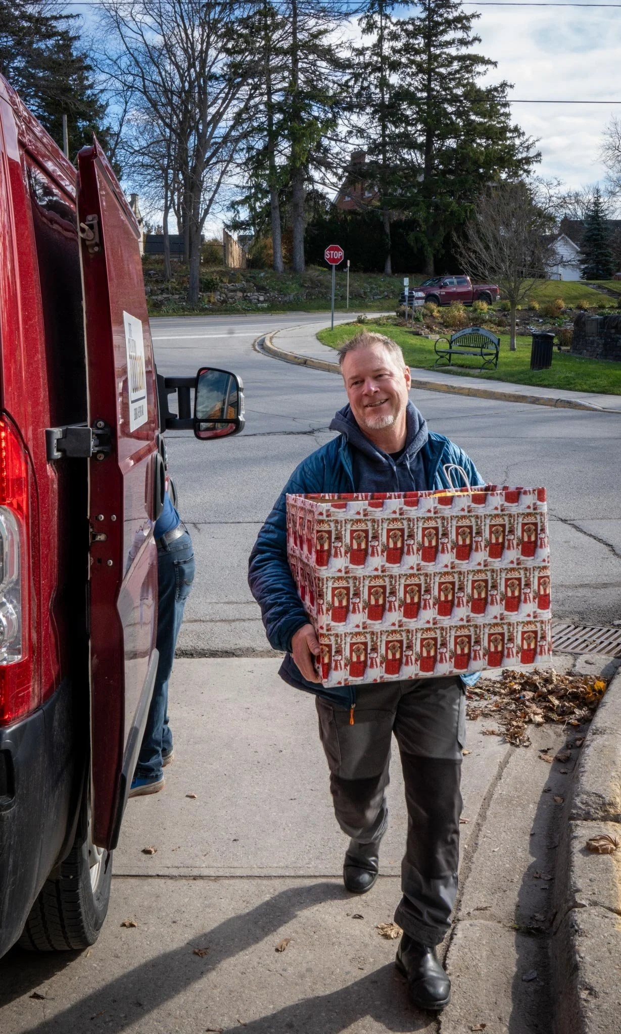 A man smiling and wearing a blue jacket carrying a large Christmas-themed gift bag, walking away from a red vehicle on a driveway in a suburban neighborhood.