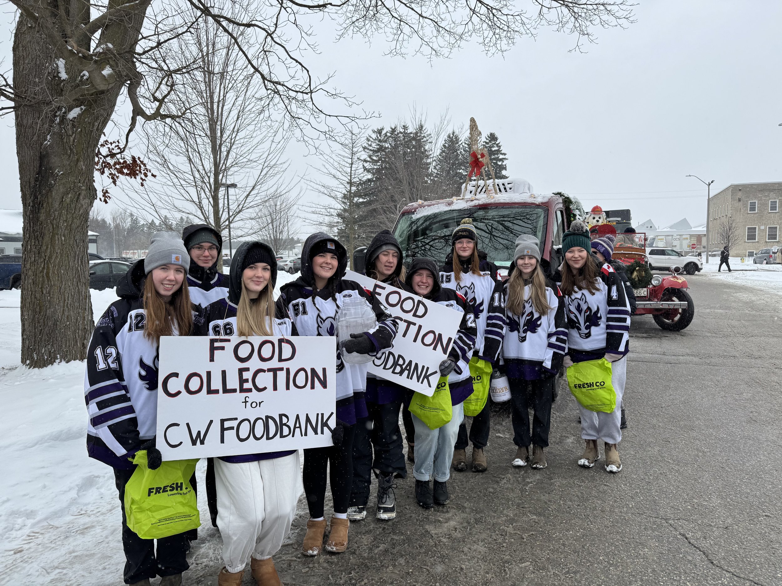 A group of young people wearing hockey jerseys and winter clothing standing on a snow-covered street, holding signs that read "FOOD COLLECTION for CW FOOD BANK," participating in a community event near a decorated vehicle with holiday decorations, including a small Christmas tree and a snowman figurine.