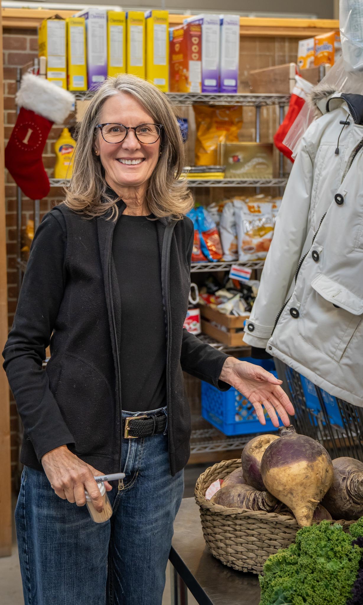 A woman with glasses and shoulder-length hair shopping in a grocery store, standing next to a basket of beets and greens, smiling at the camera.